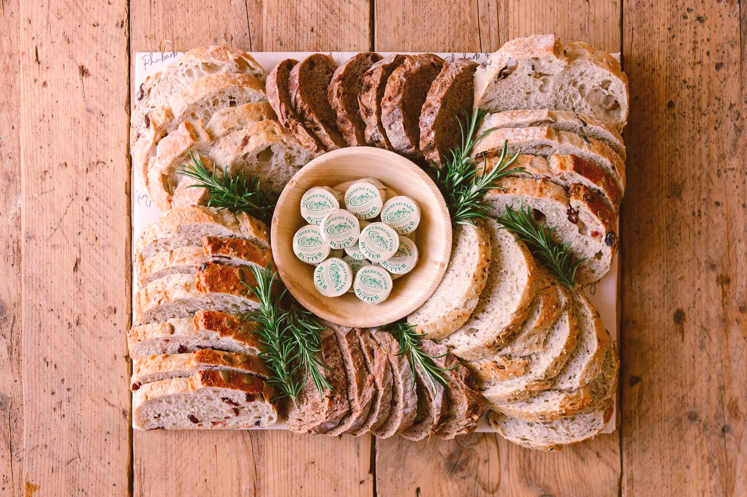 A variety of sliced breads with different textures and flavors arranged on a wooden tray, garnished with sprigs of rosemary, and surrounded by a circle of butter pats in a wooden dish on a rustic wooden table.