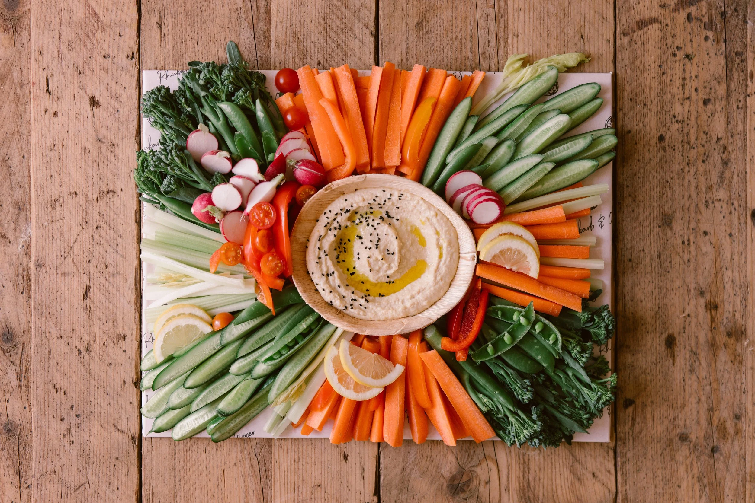 A platter of assorted fresh vegetables including carrots, cherry tomatoes, radishes, cucumber, celery, and bell peppers, with a central bowl of hummus garnished with black sesame seeds and olive oil, on a wooden table.