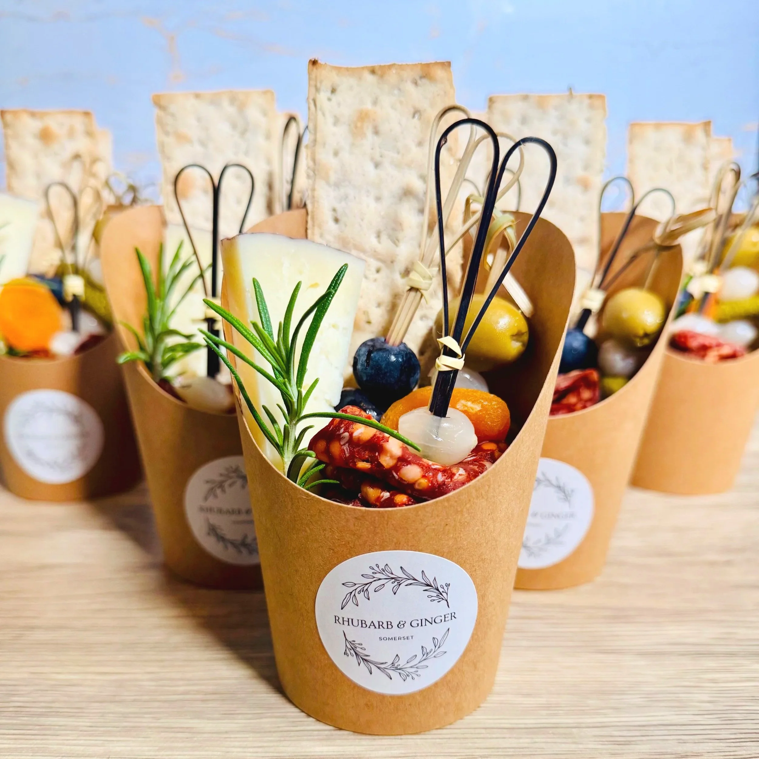 Sandwiches served in brown paper cups with cheese, crackers, fresh herbs, fruit, nuts, and skewers on a wooden surface.