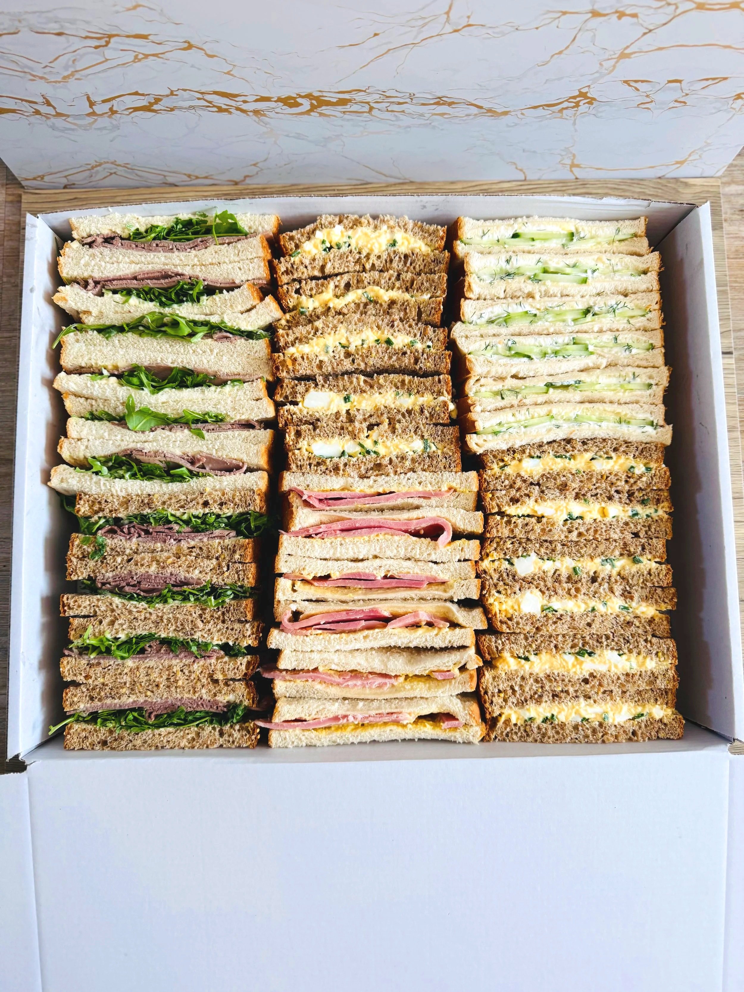 A box of assorted sandwiches with three varieties: with greens and roast beef, with egg salad, and with cucumber and dill on whole wheat, held in a white box.