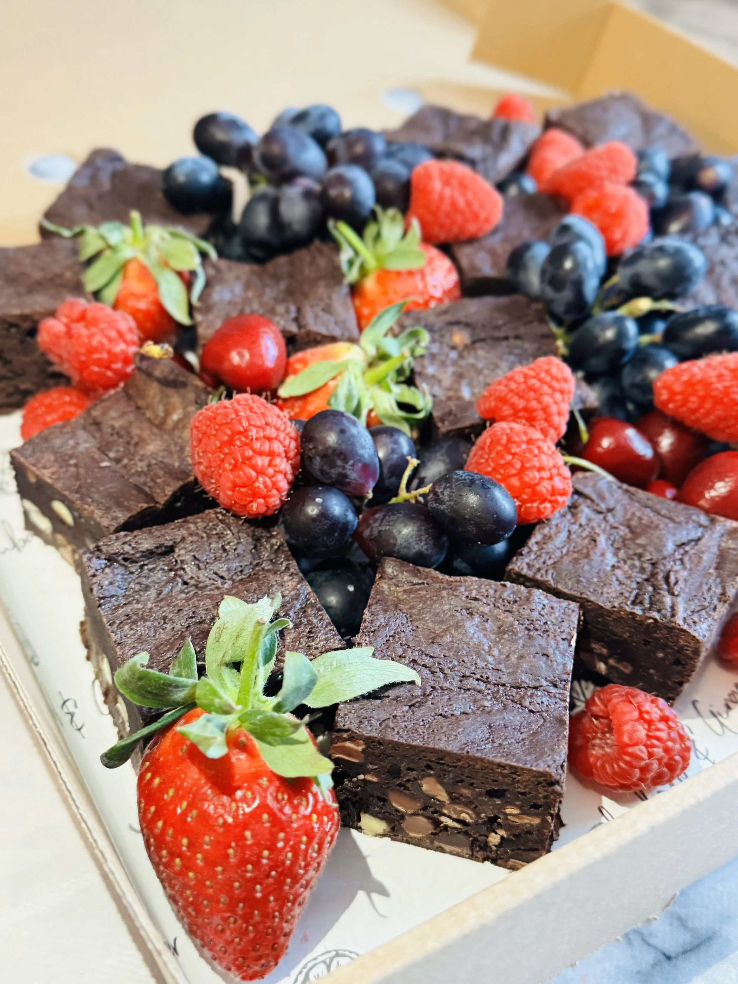 Close-up of a box of chocolate brownie squares topped with fresh strawberries, blackberries, and raspberries.