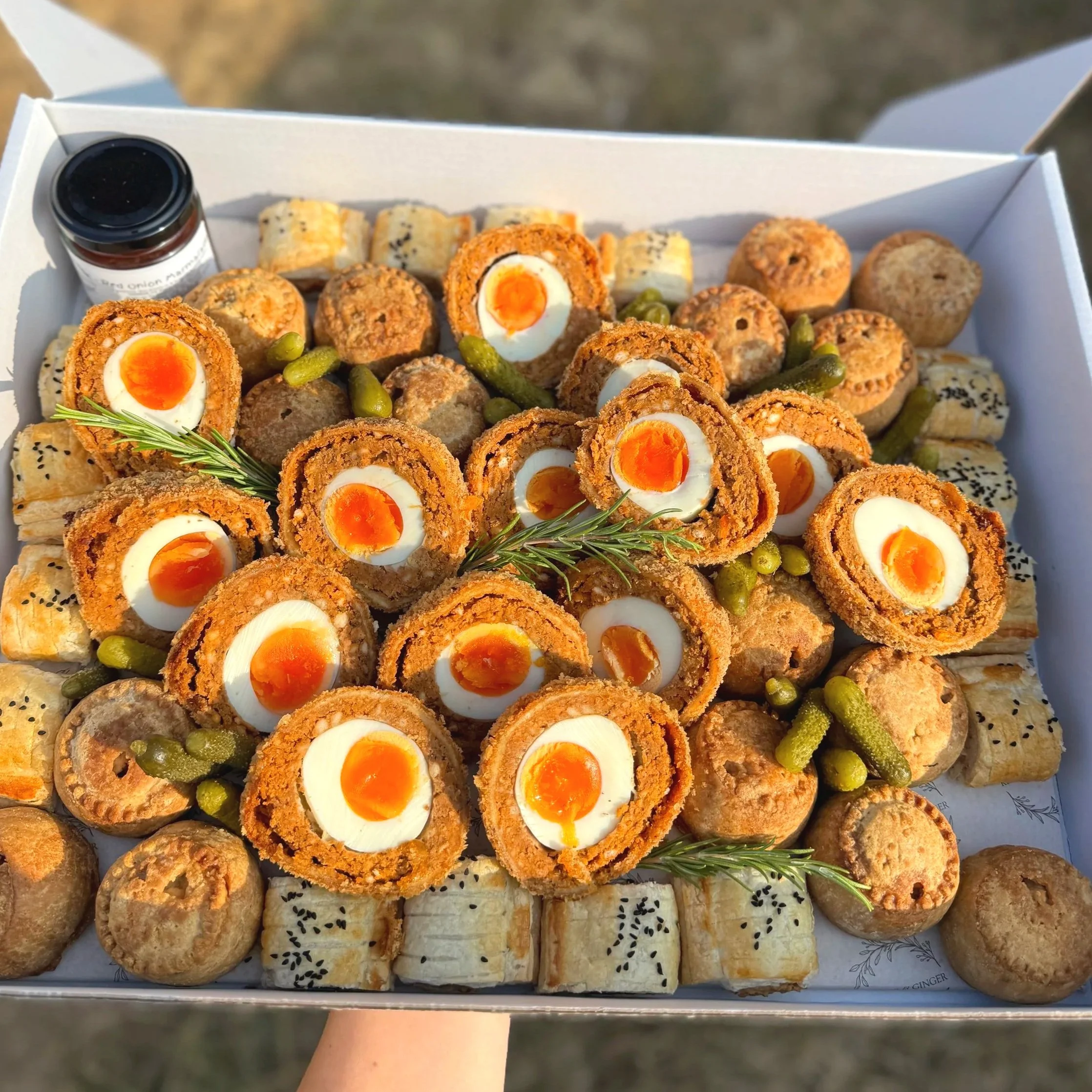A tray of assorted appetizers including Scotch eggs with soft boiled eggs inside, small savory pastries topped with black sesame seeds or black seeds, and round, pastry-like bites garnished with rosemary sprigs and green pickles.