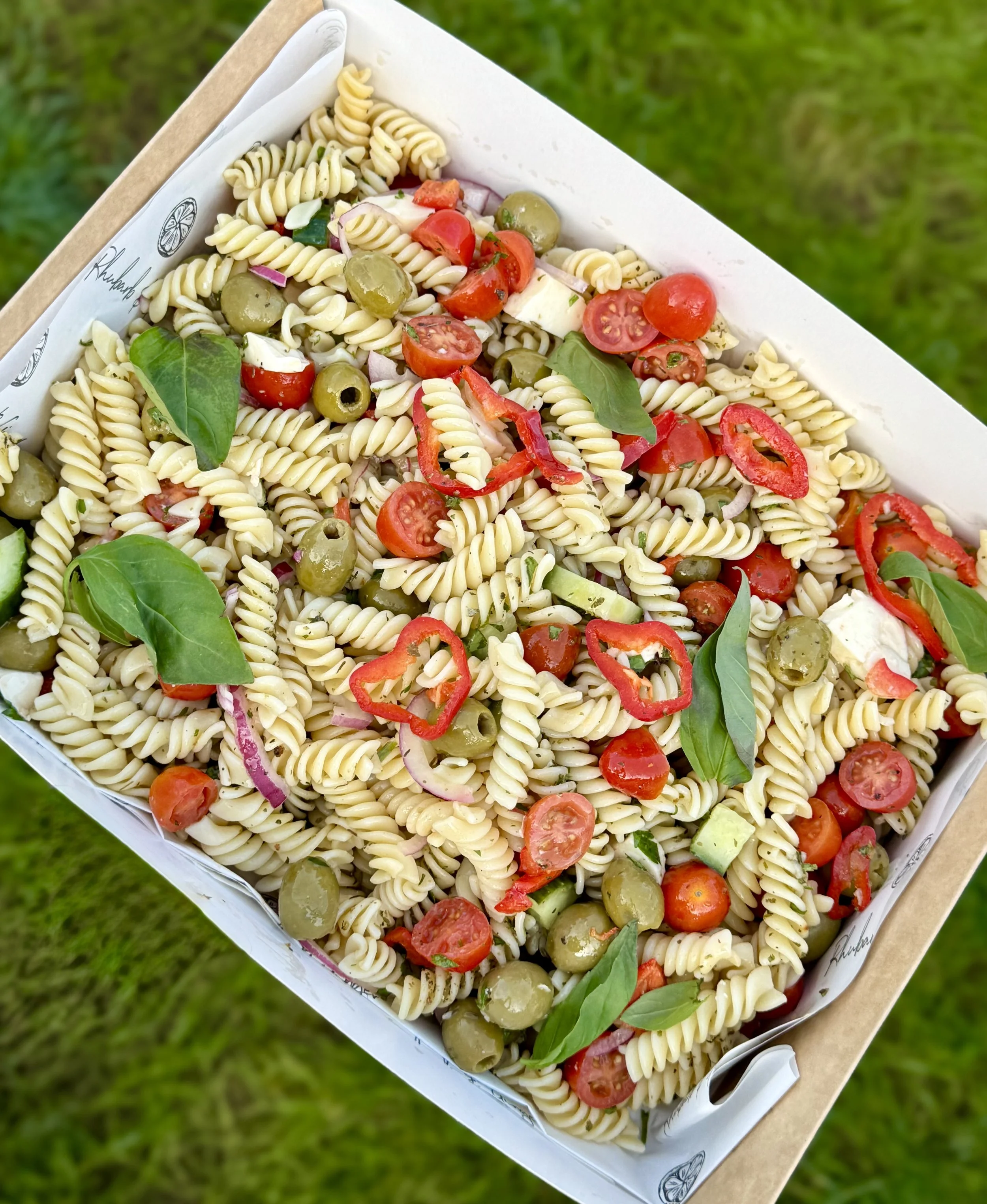 Fresh pasta salad with cherry tomatoes, green olives, red bell pepper slices, cucumber, red onion, and basil leaves in a takeout container.
