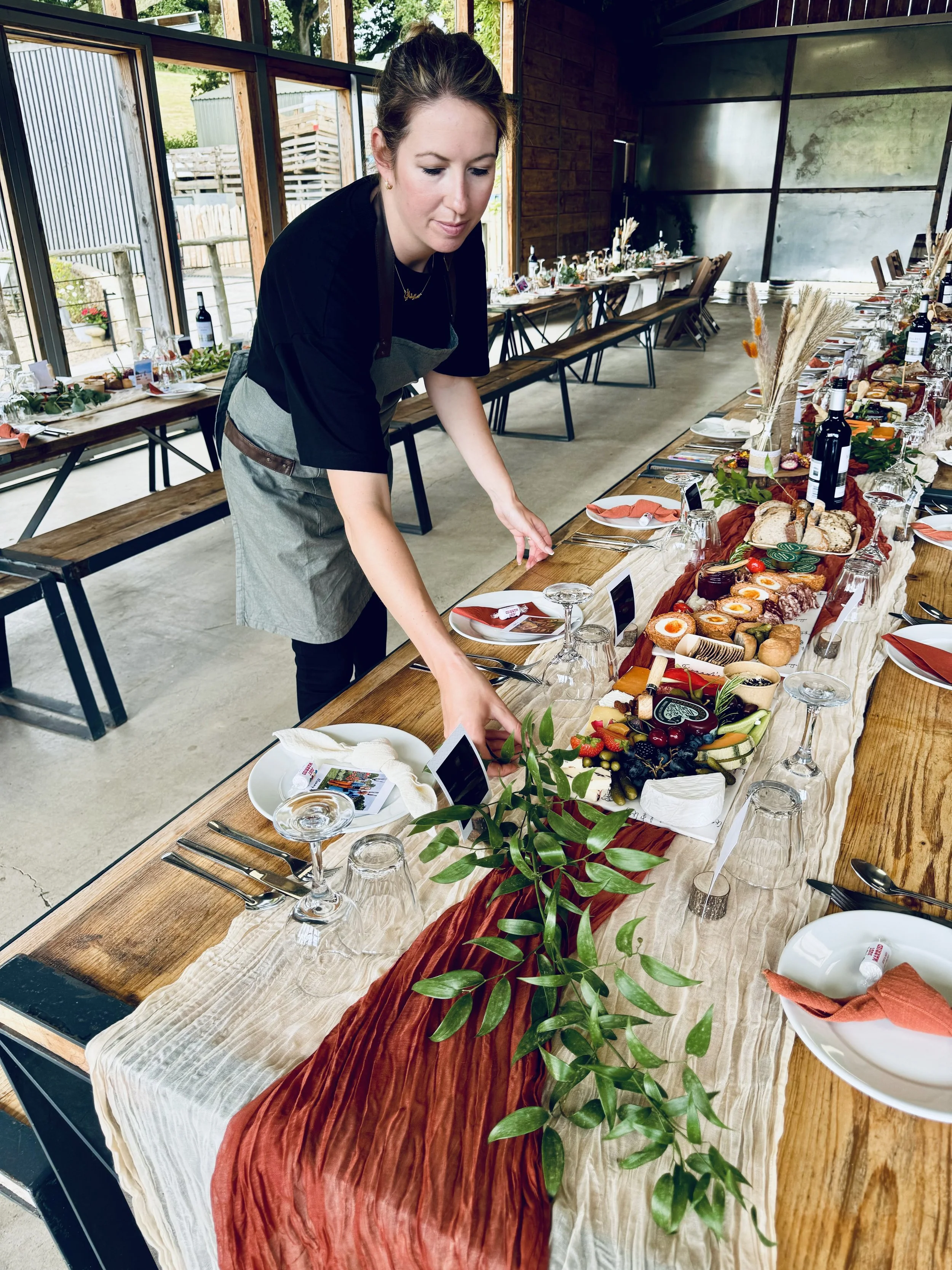 A woman in a black shirt and gray apron arranging a long, decorated dining table filled with assorted foods, drinks, and table settings in a rustic indoor space with large windows.
