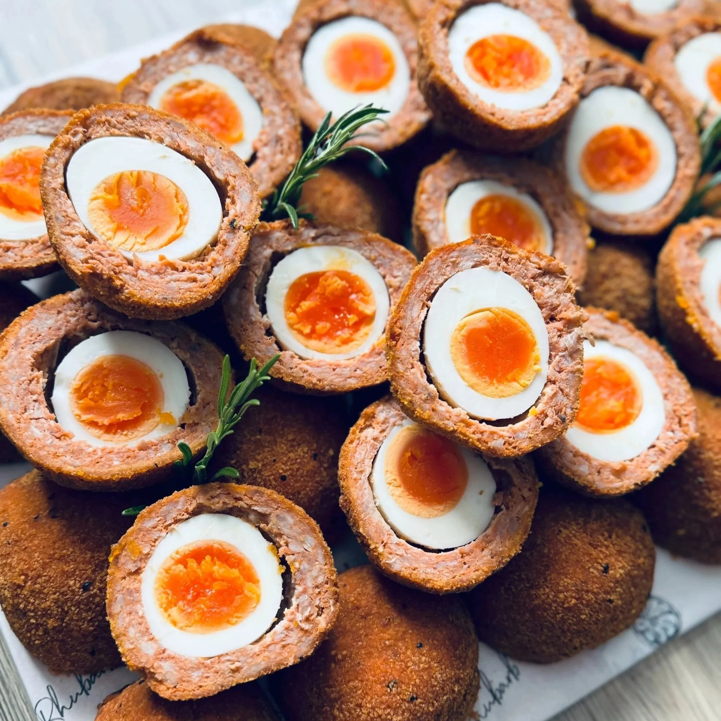 Close-up of Scotch eggs, halved to show hard-boiled egg and sausage meat, garnished with rosemary sprigs, on a plate