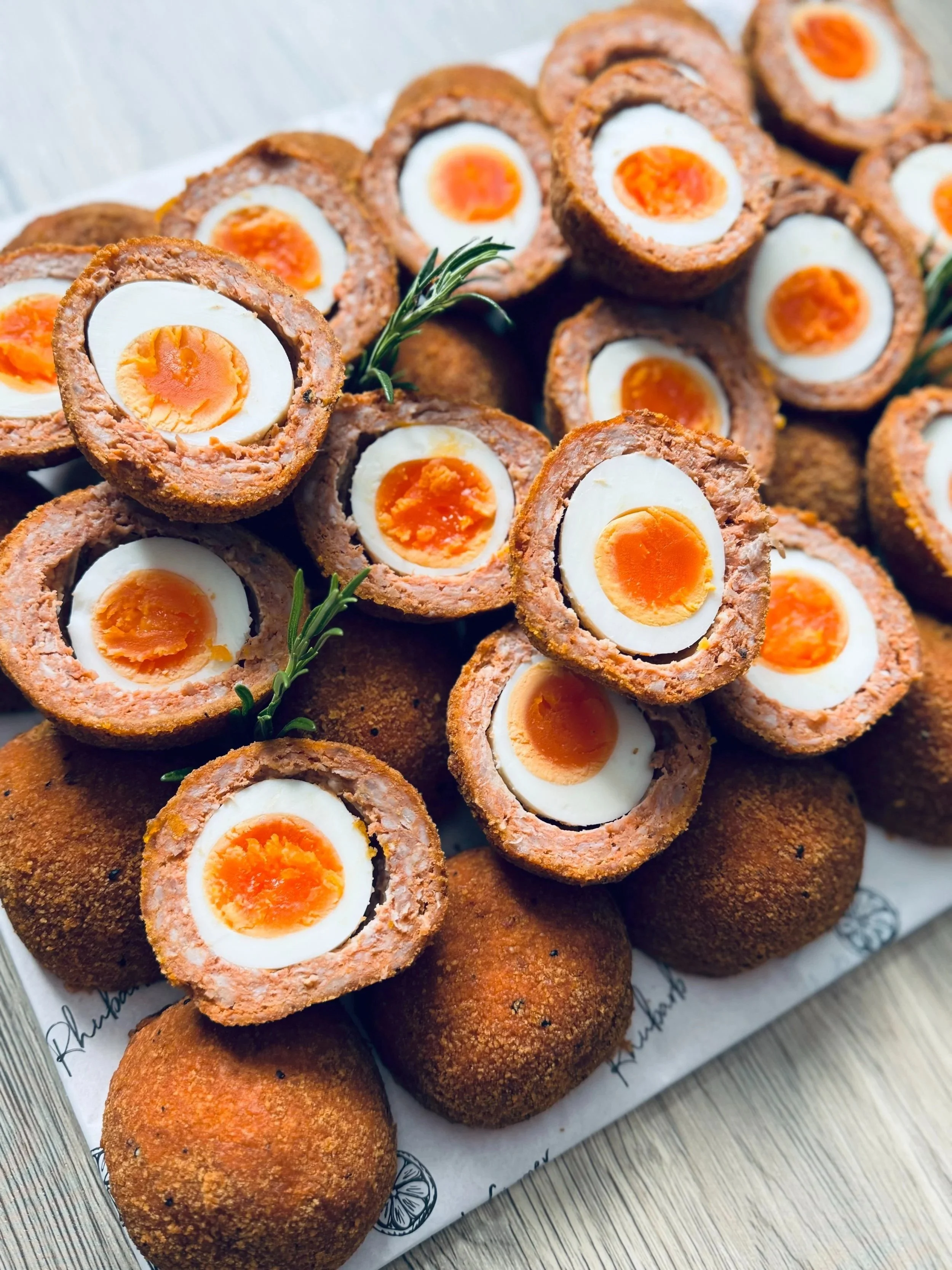 Close-up of Scotch eggs and falafel balls on a wooden surface, garnished with sprigs of rosemary.