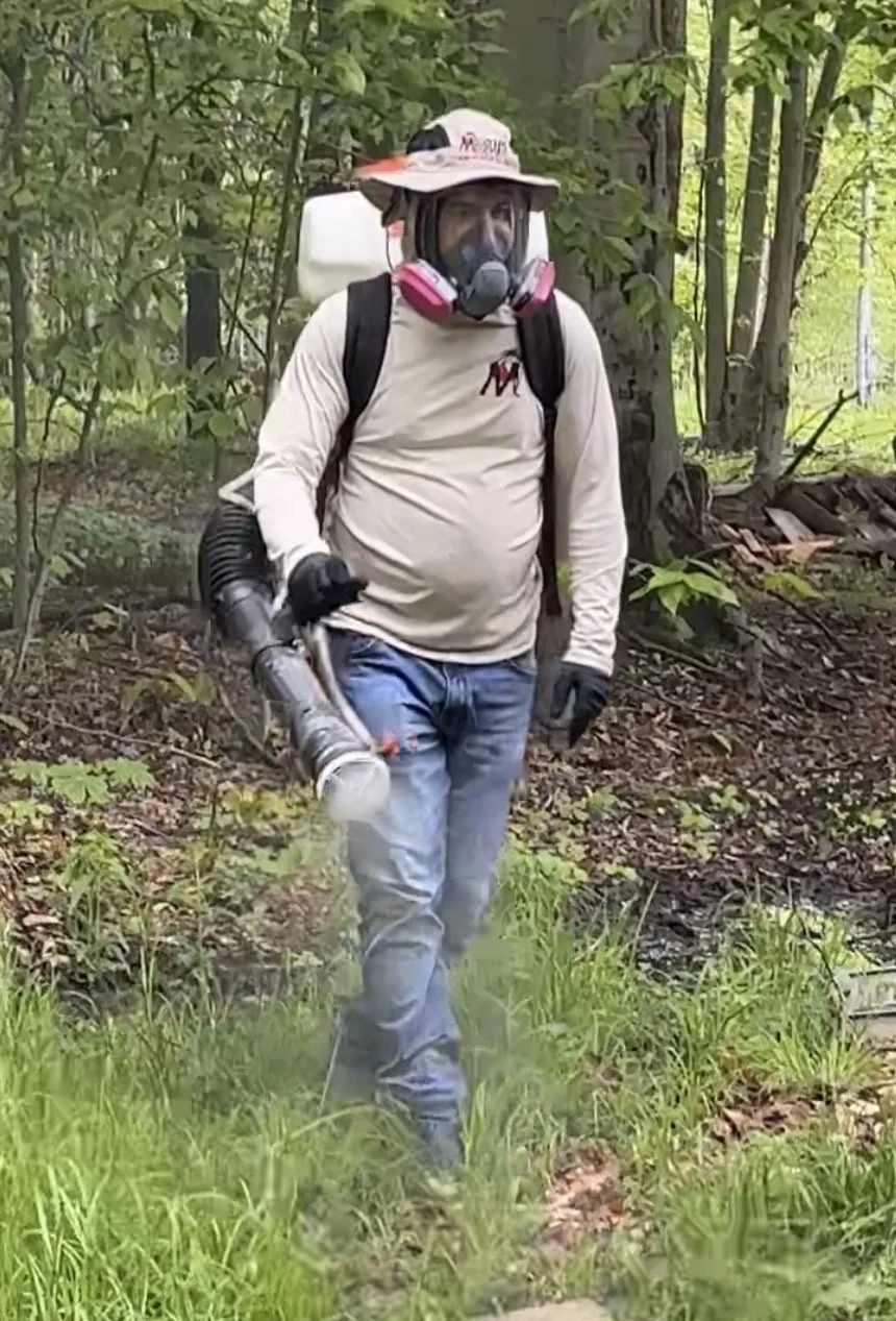 Mosquito Assassin Pest Control technician applying mosquito treatment outdoors while wearing protective equipment and backpack sprayer.