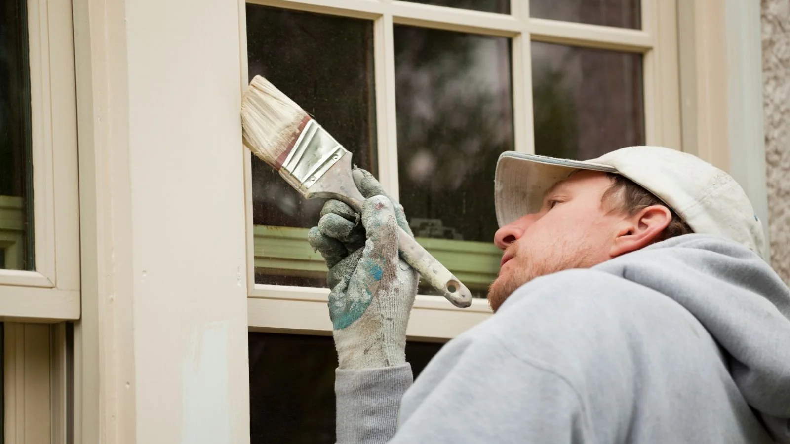 A man wearing a gray hoodie, gloves, and a white cap paints the trim of a window with a paintbrush.