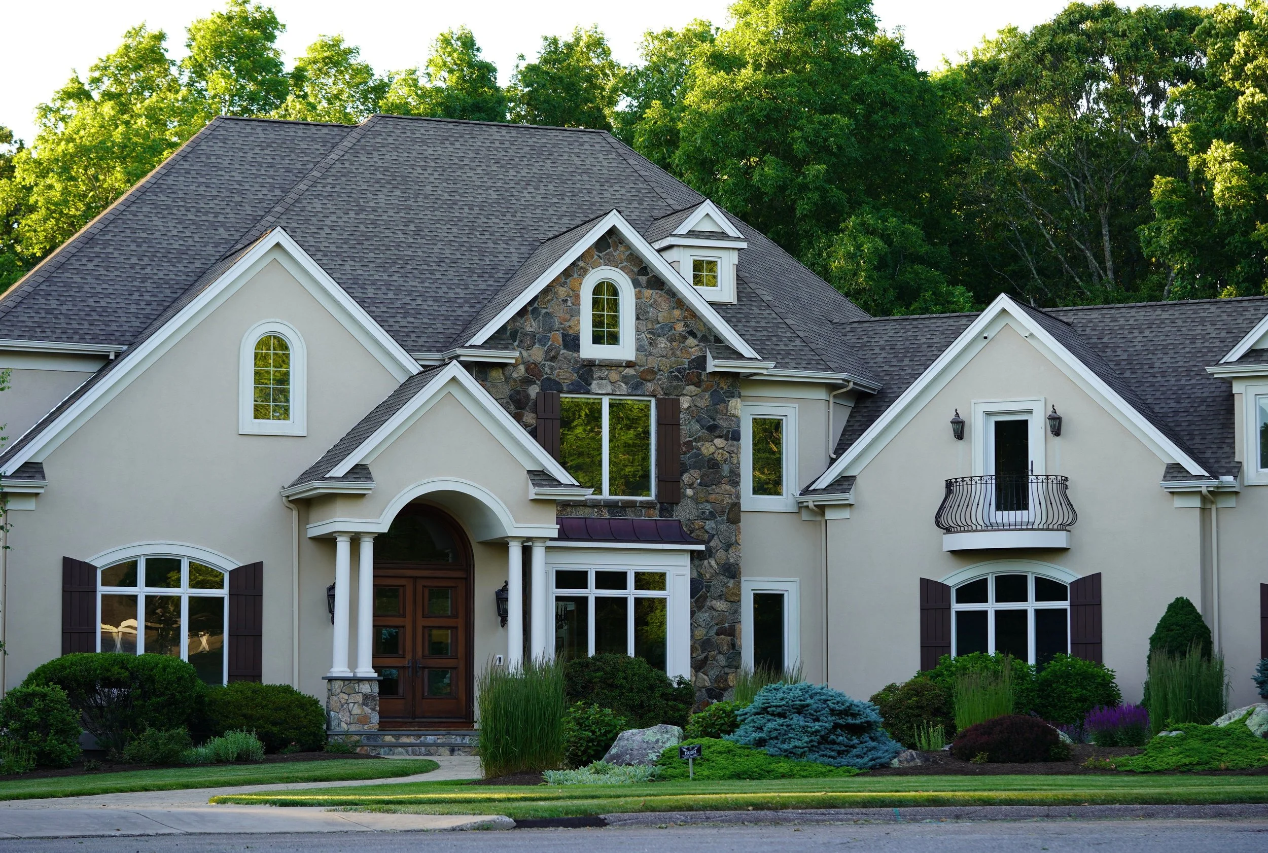 Front view of a large suburban house with a stone and stucco exterior, multiple gables with white trim, arched windows, a small balcony, and lush landscaped garden in the foreground, surrounded by tall green trees.
