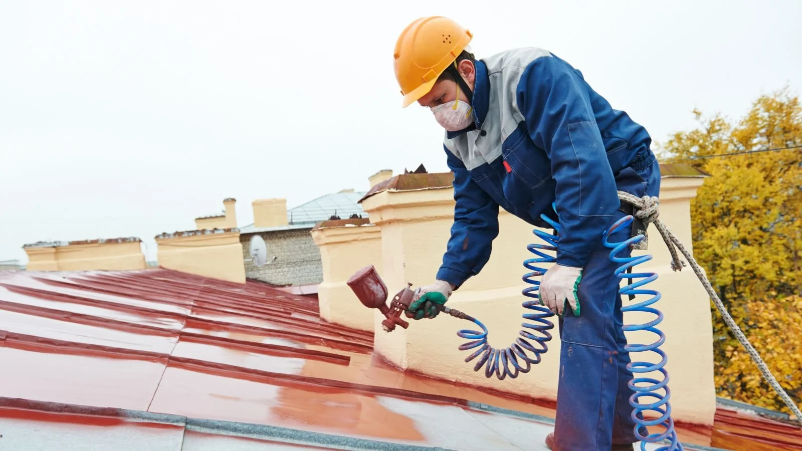 A worker wearing a yellow safety helmet, face mask, gloves, and a blue work uniform is spray painting a red metal roof using a spray gun connected to a blue hose.
