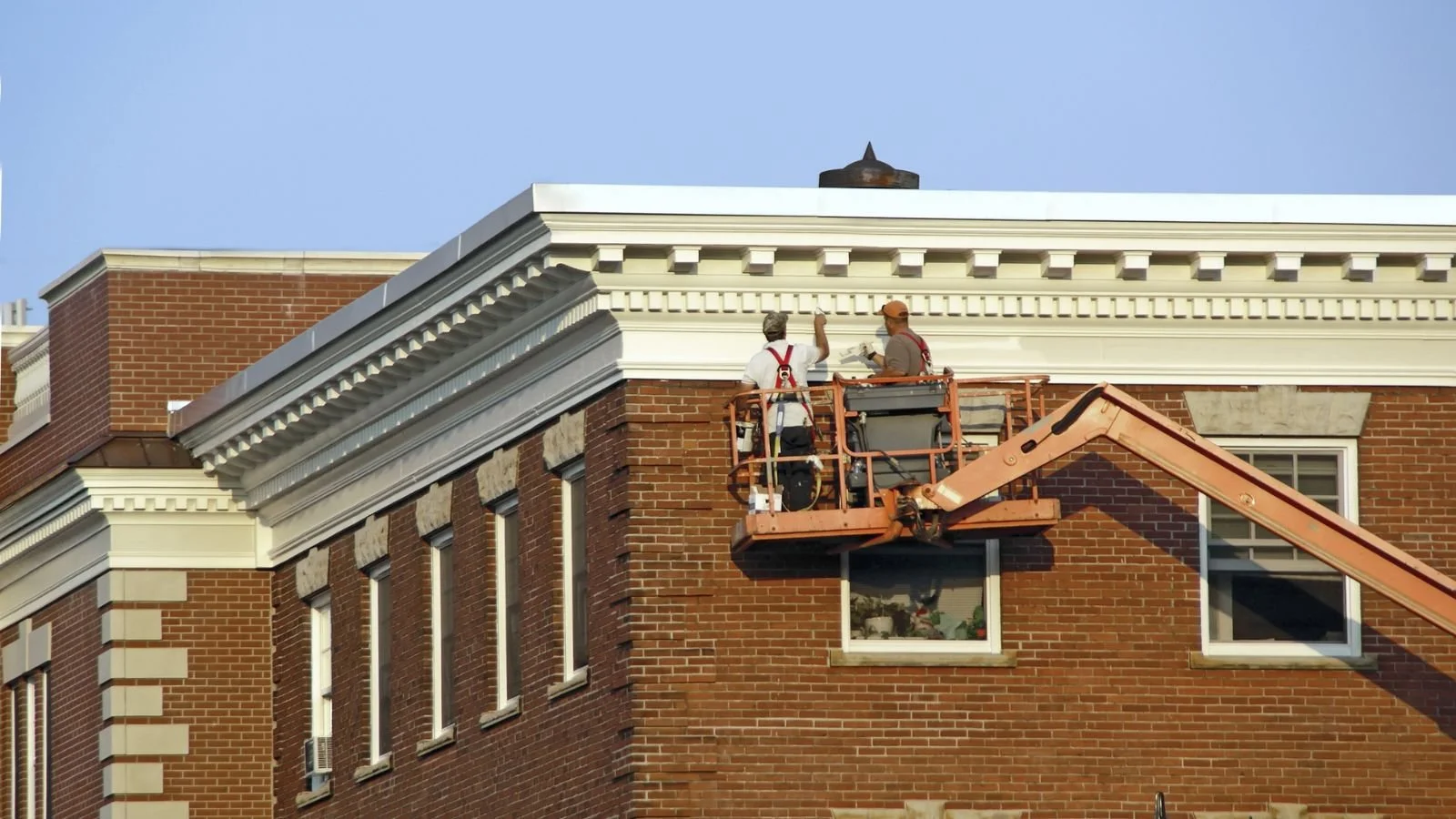 Two construction workers in orange safety gear on an elevated lift working on the corner of a brick building with white decorative molding under a clear blue sky.
