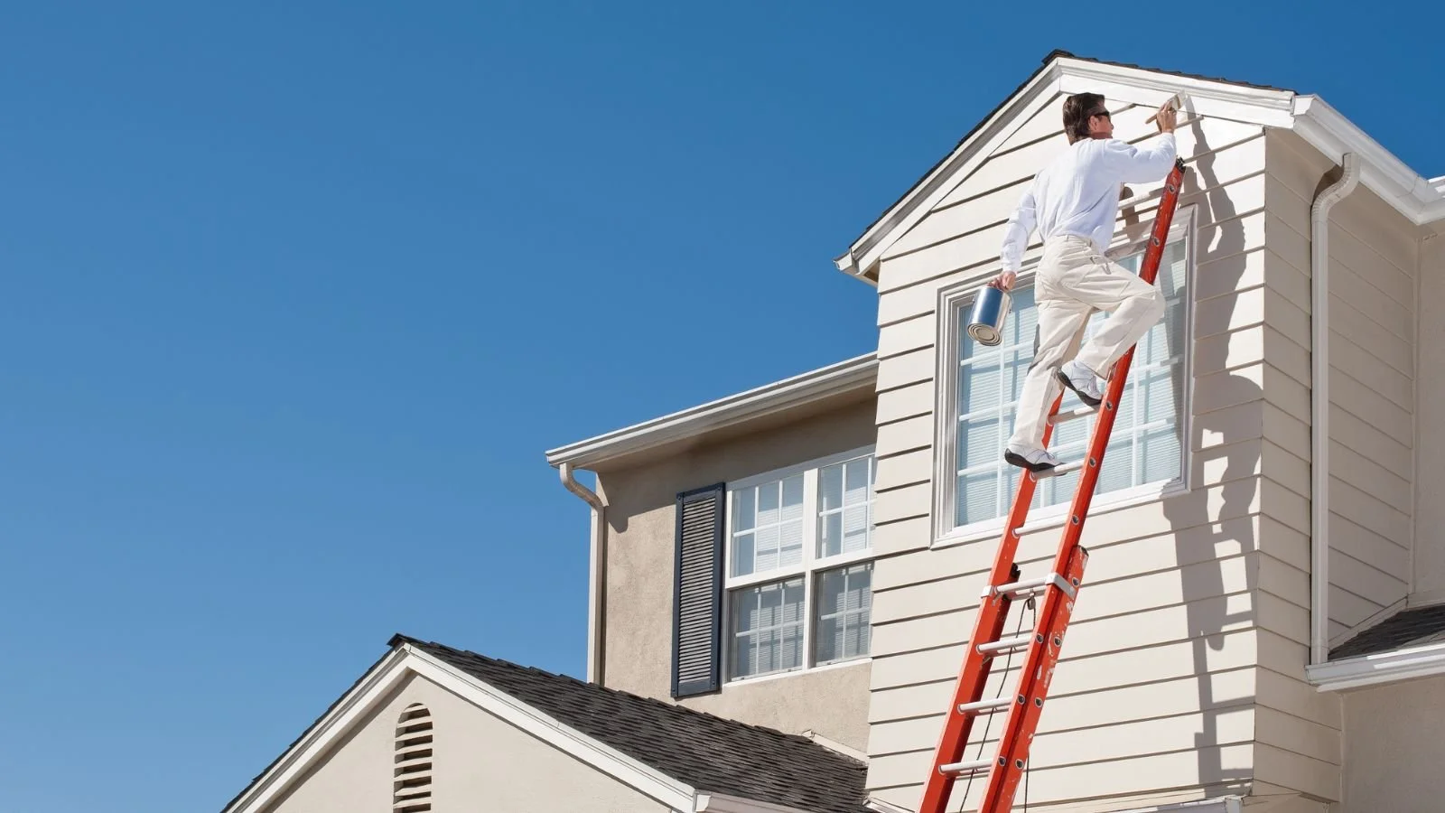 A man in a white shirt and beige pants painting the exterior wall of a house from a tall orange ladder, holding a roller brush and a can of paint under a clear blue sky.