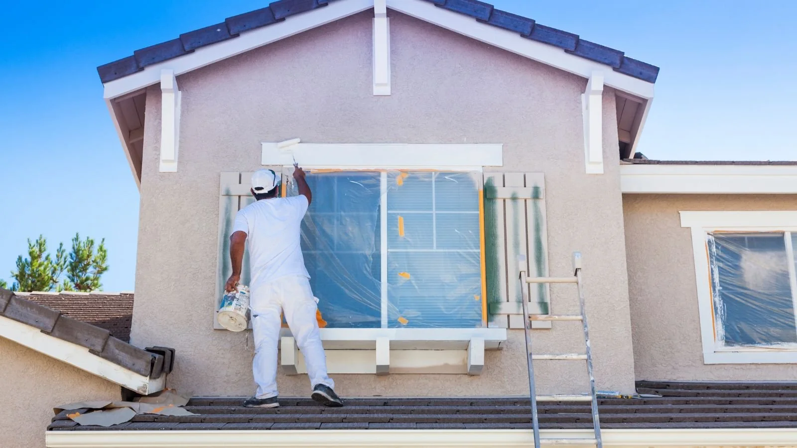 A man painting the trim of a large window on the second floor of a house with beige stucco exterior. The window is covered with plastic for protection. A ladder is set against the house on the right, and a paint bucket is in the man's hand. The roof is brown, and the sky is clear and blue.