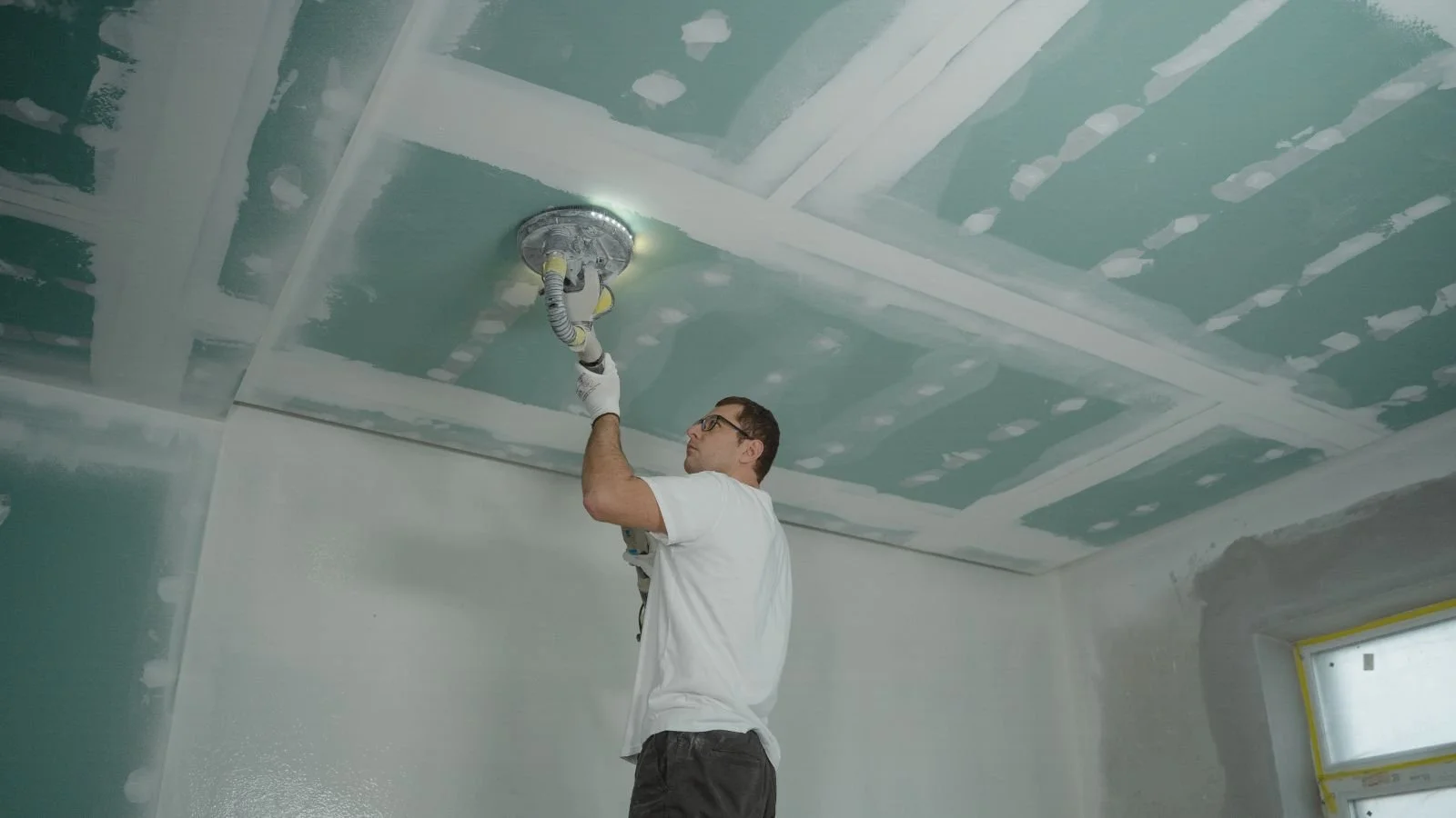 A man wearing glasses and a white t-shirt is sanding or smoothing a ceiling with a power tool, inside a room under construction or renovation.