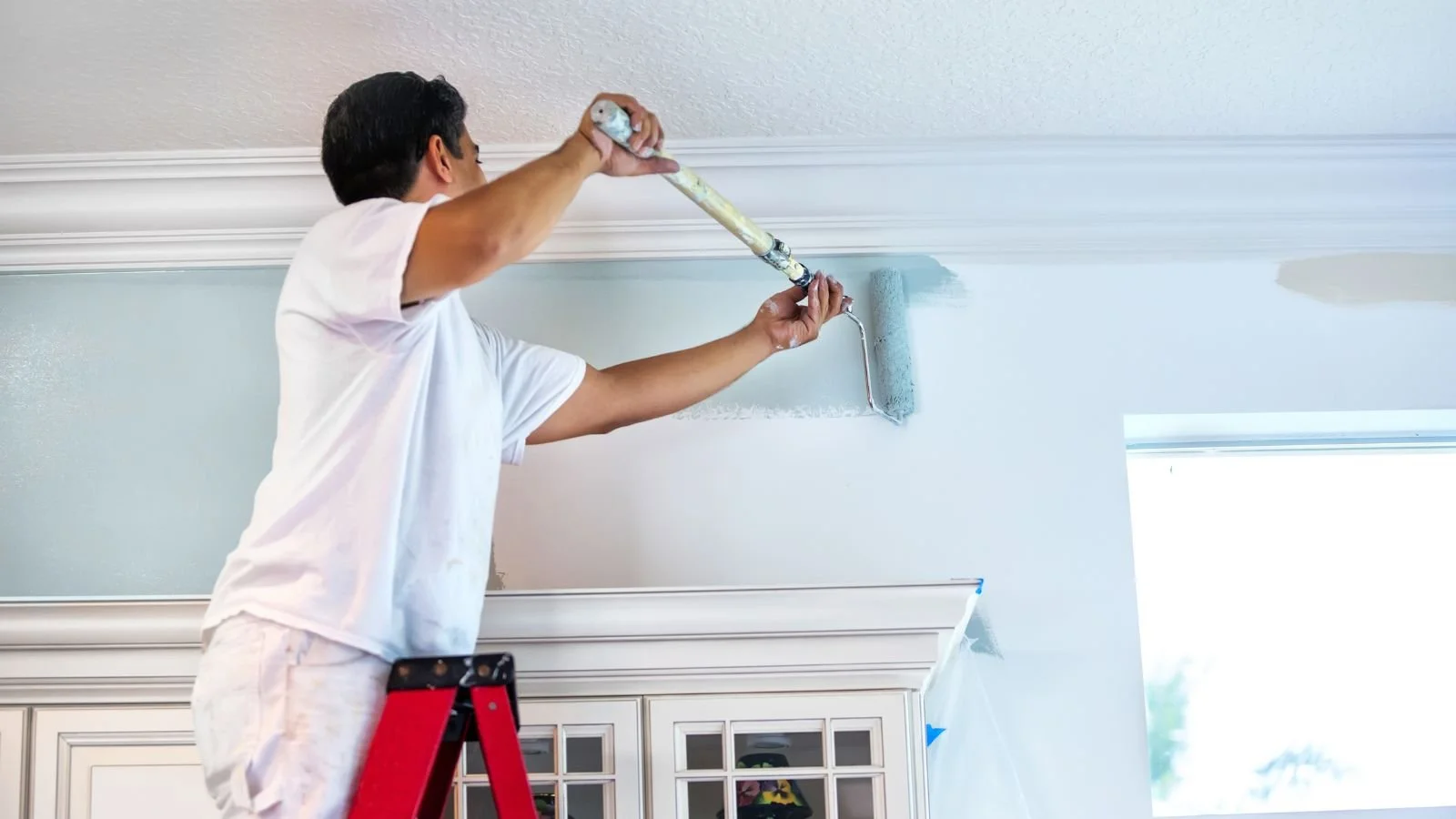 A man painting a ceiling with a paint roller while standing on a ladder in a living room.