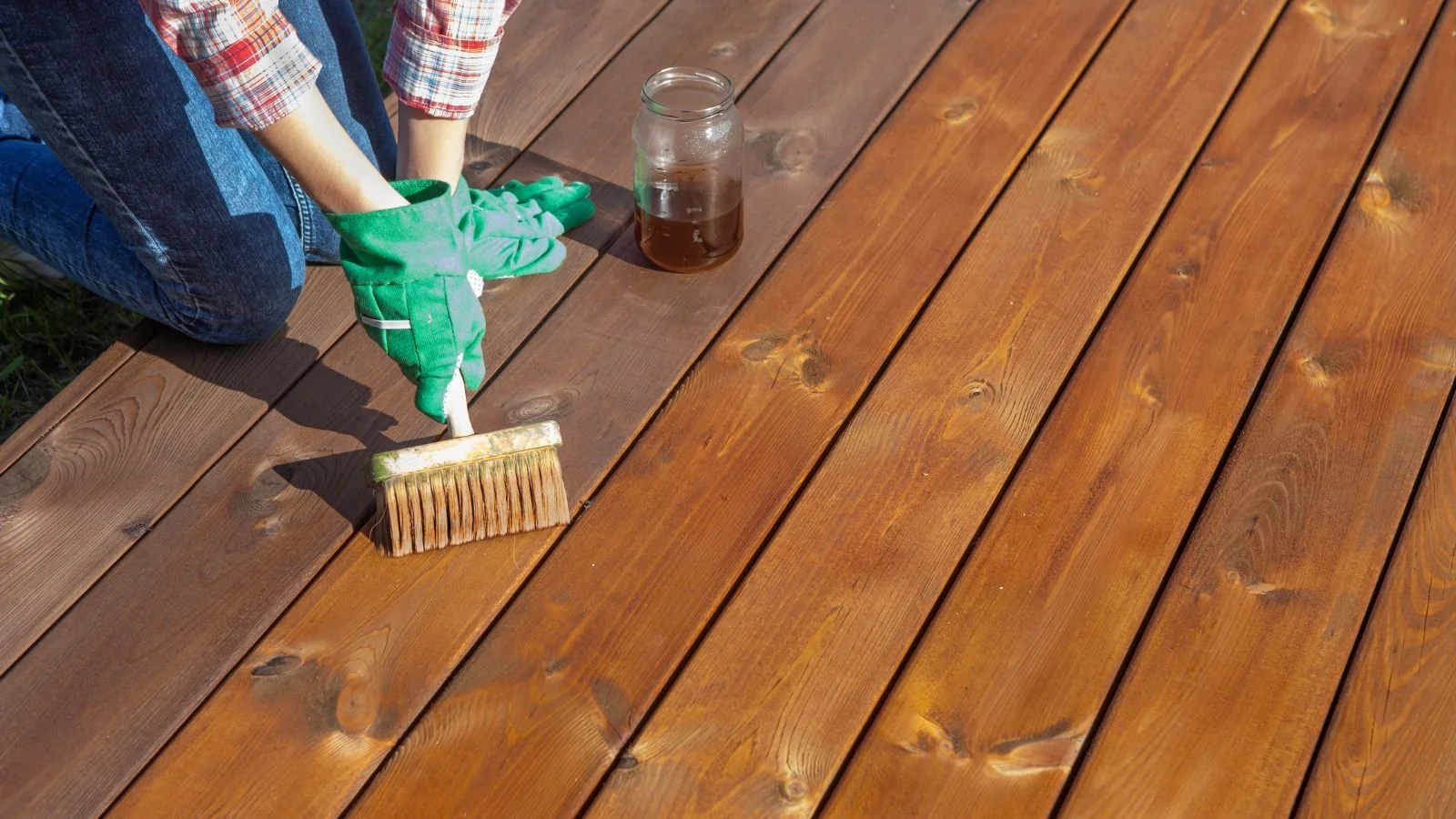 Person in plaid shirt and jeans applying a stain or finish to a wooden deck with a brush, while kneeling on the deck. There is a jar of stain nearby.