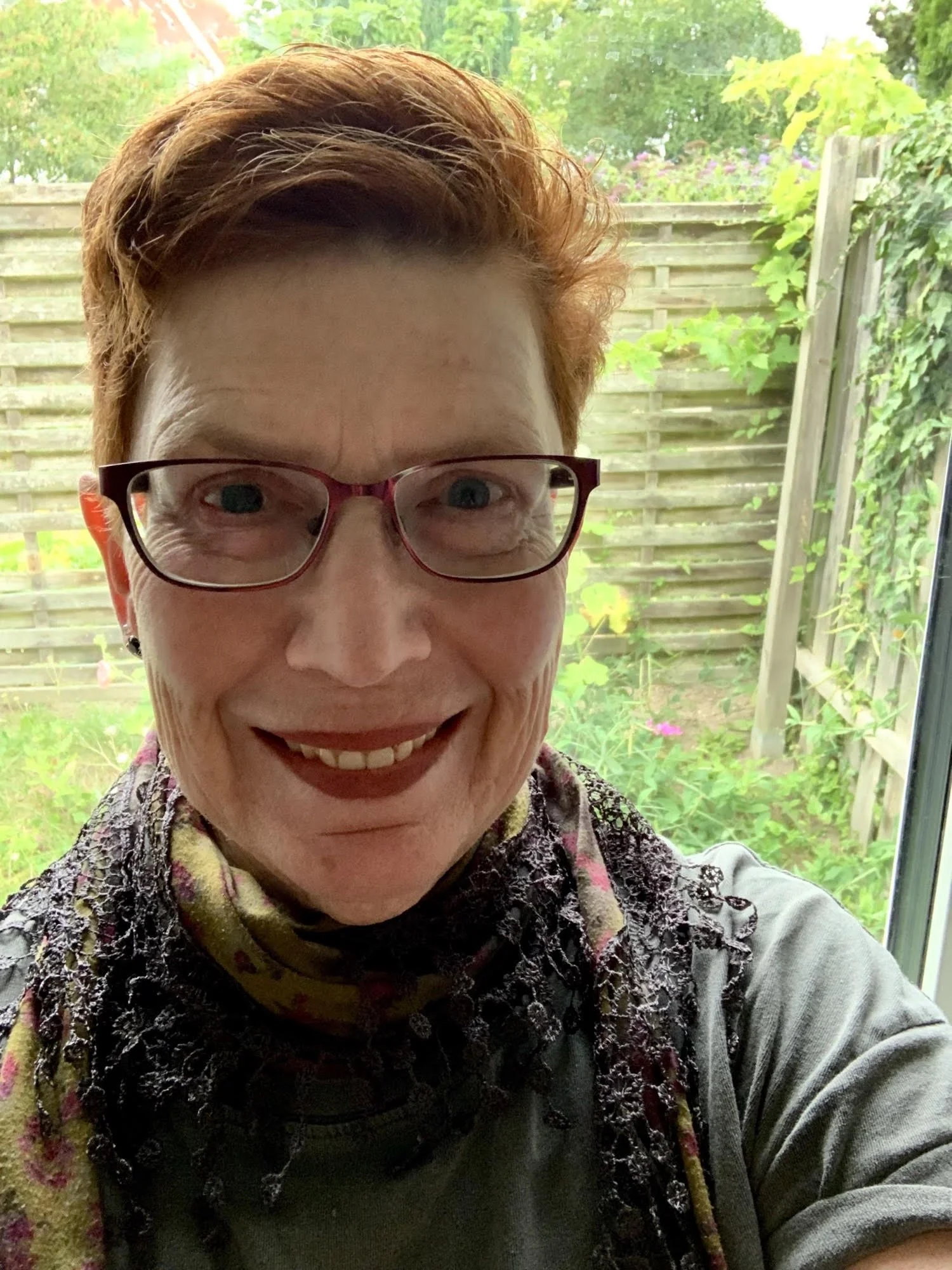 A smiling woman with short red hair and glasses taking a selfie indoors near a window with a view of a backyard garden with trees and wooden fence.