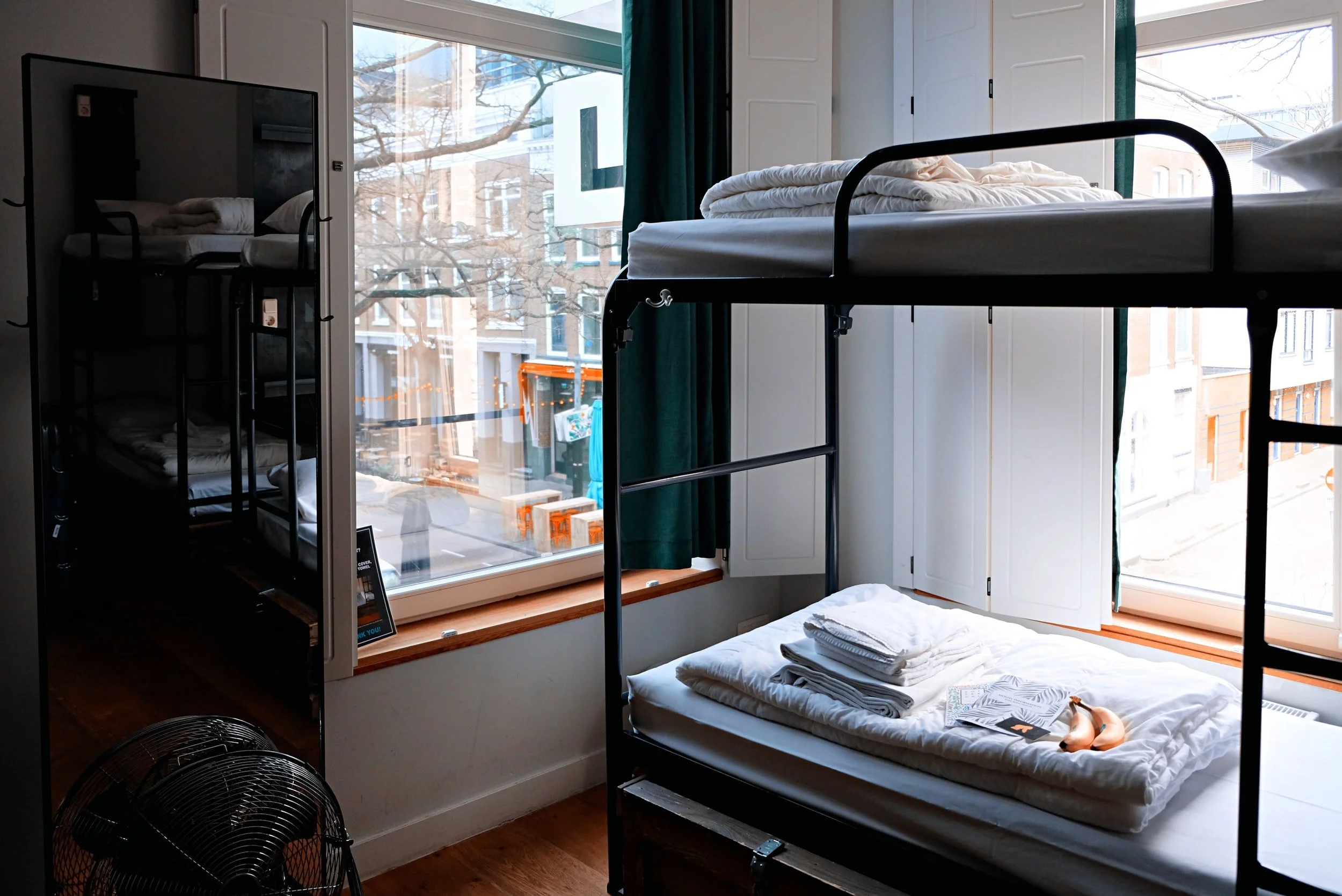 Bedroom with bunk beds, large windows, and a reflection in a mirror. The bed has folded linens and a banana on top. Outside view shows urban buildings.