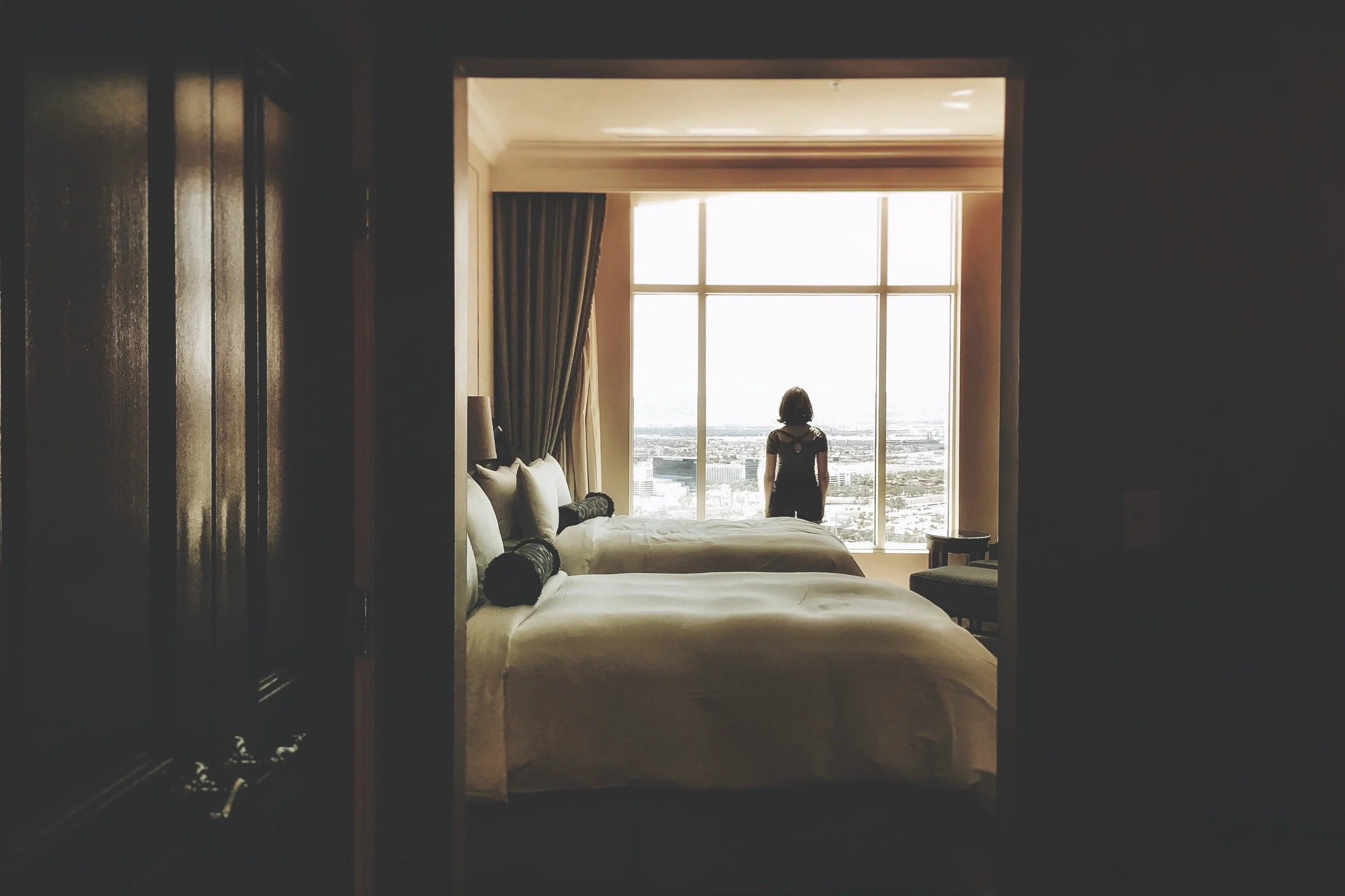 Interior of a hotel room with a person standing by a large window, overlooking the cityscape. The room features two made beds and curtains.
