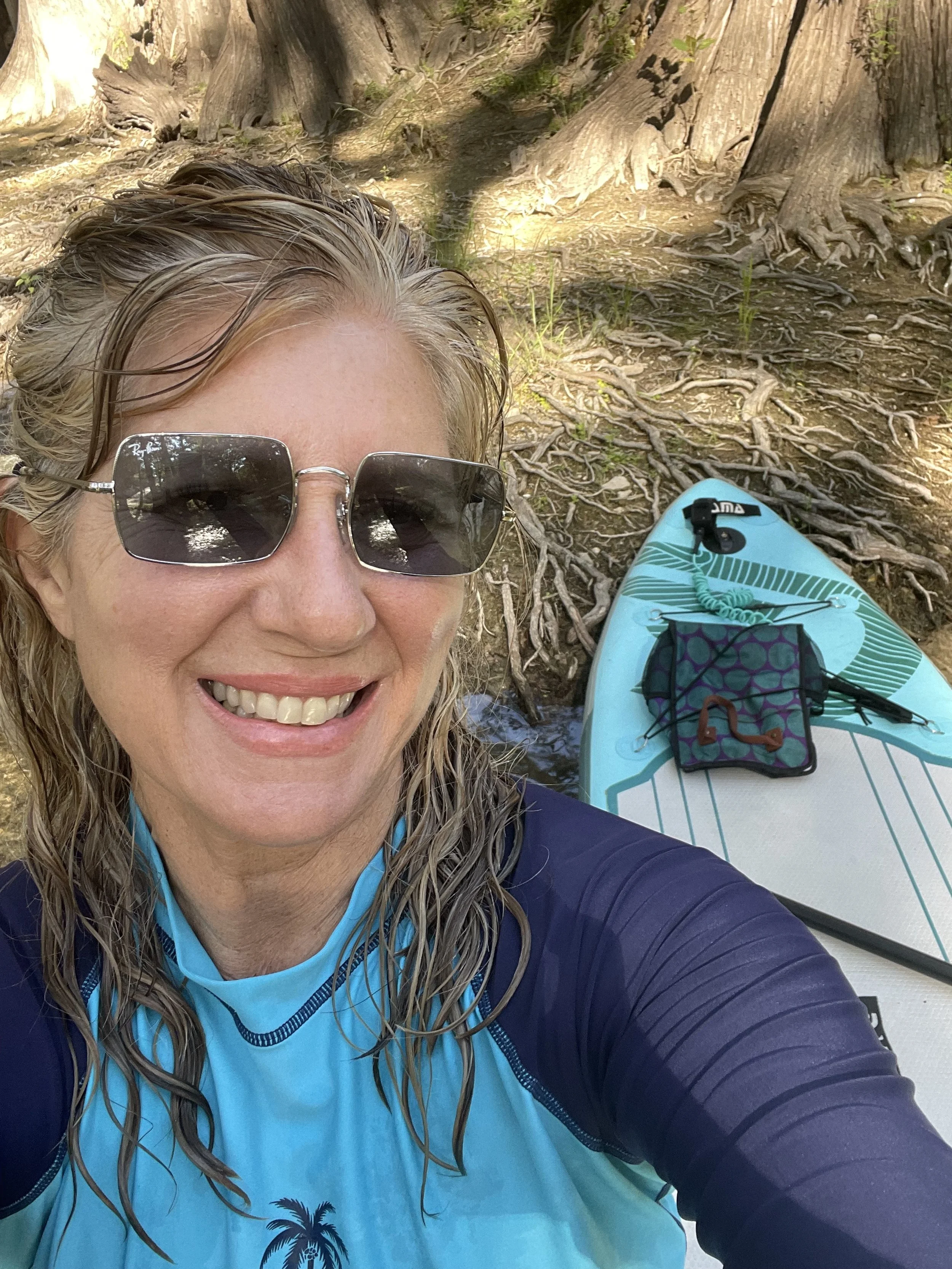 A woman with blonde, wet hair, wearing sunglasses and a blue athletic shirt, smiling for a selfie next to a paddleboard with some small items on it, in a wooded area with large tree roots.