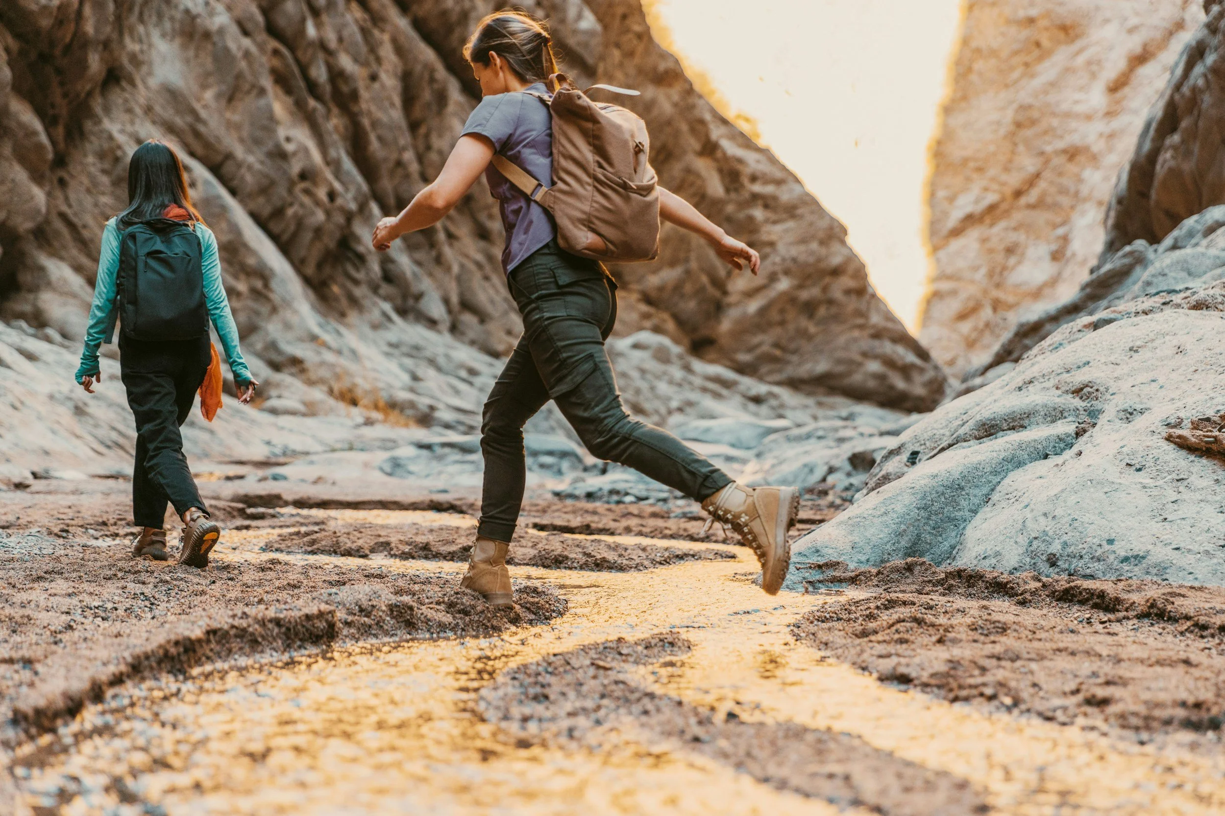 Two women hiking through a rocky canyon, wearing backpacks and hiking boots, during sunset or sunrise.