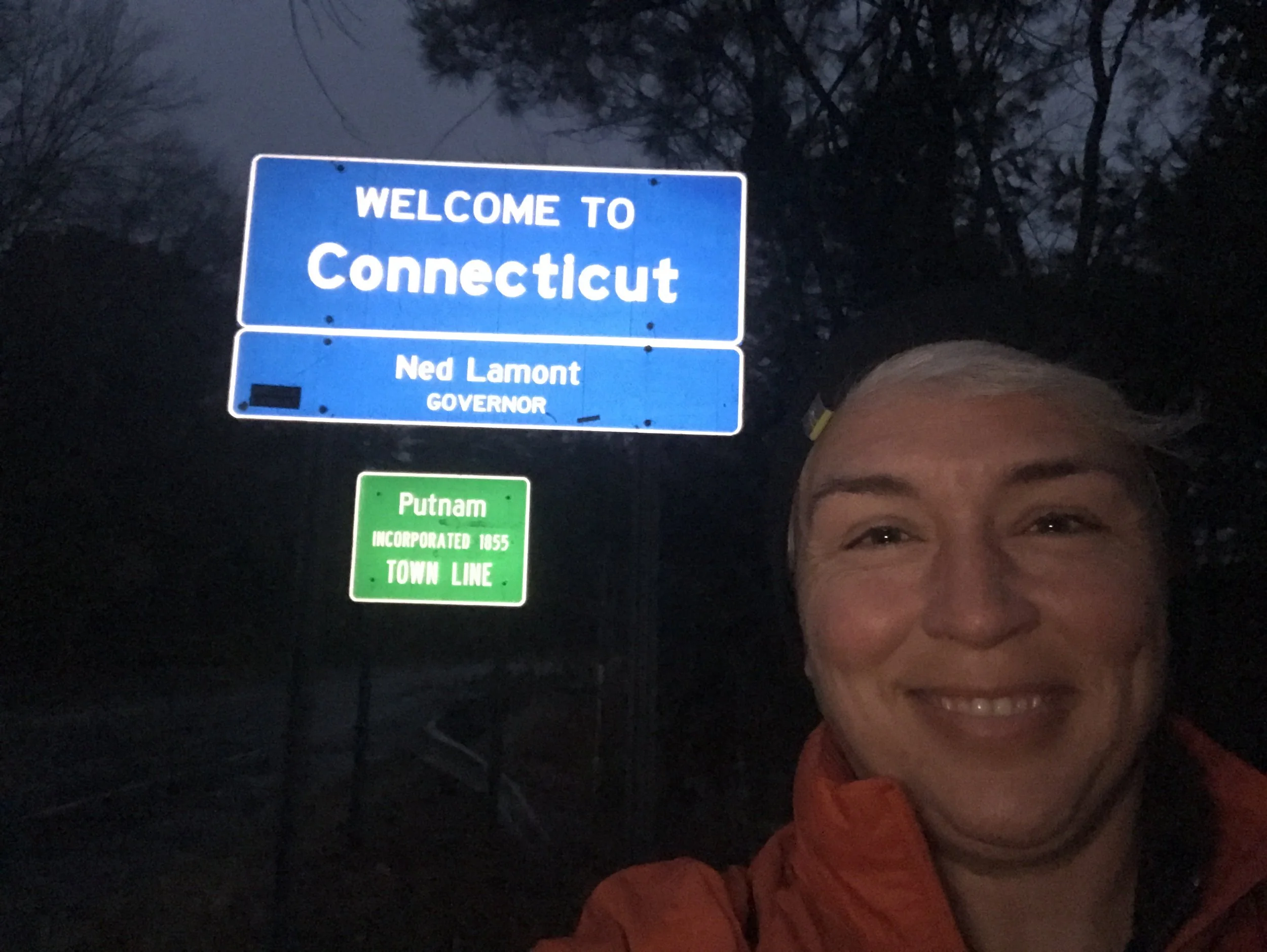 Person taking a selfie beside a "Welcome to Connecticut" sign at the Putnam town line during nighttime.