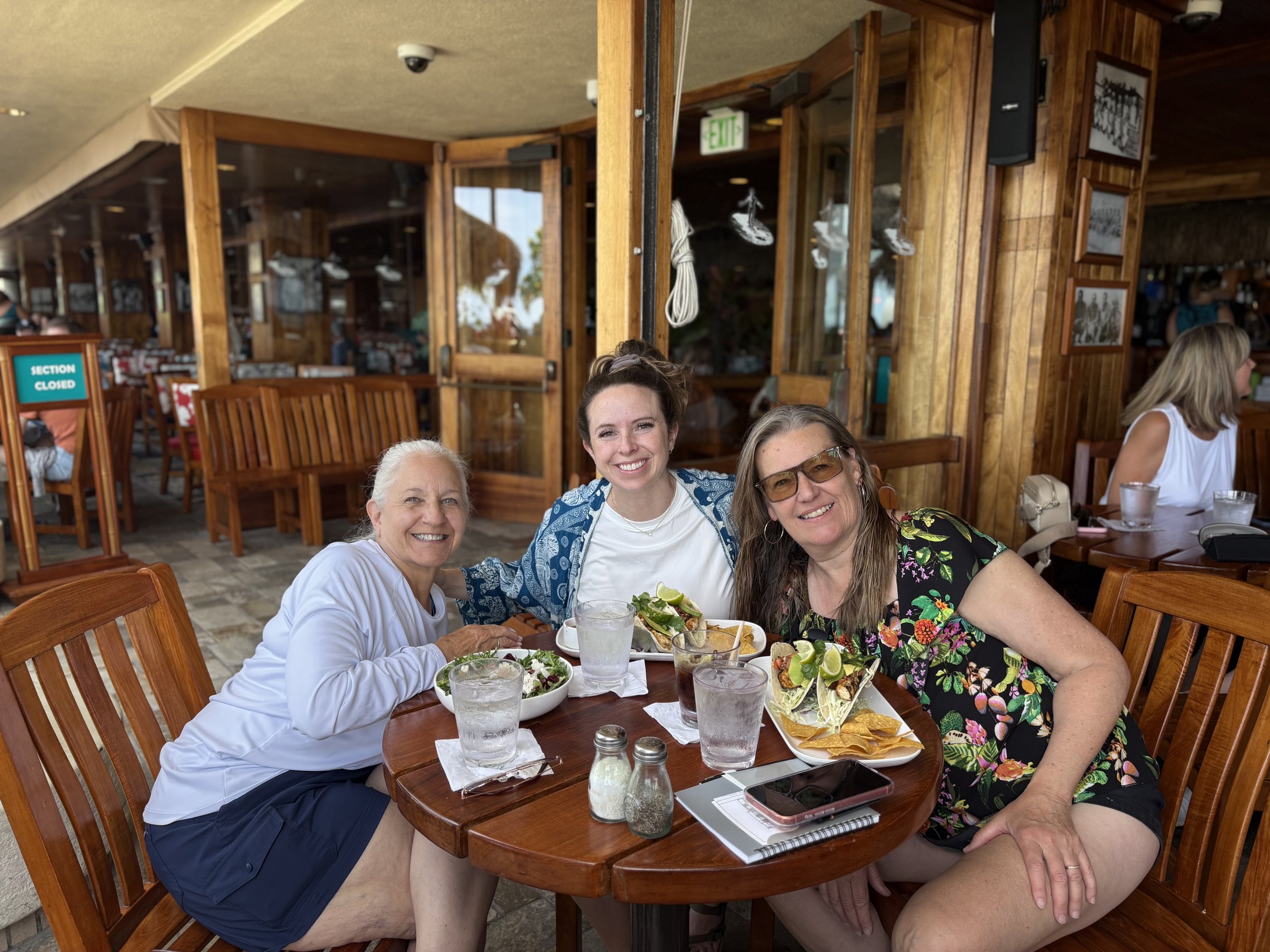 Four women sitting at a wooden table at a restaurant, smiling at the camera, with food and drinks in front of them.
