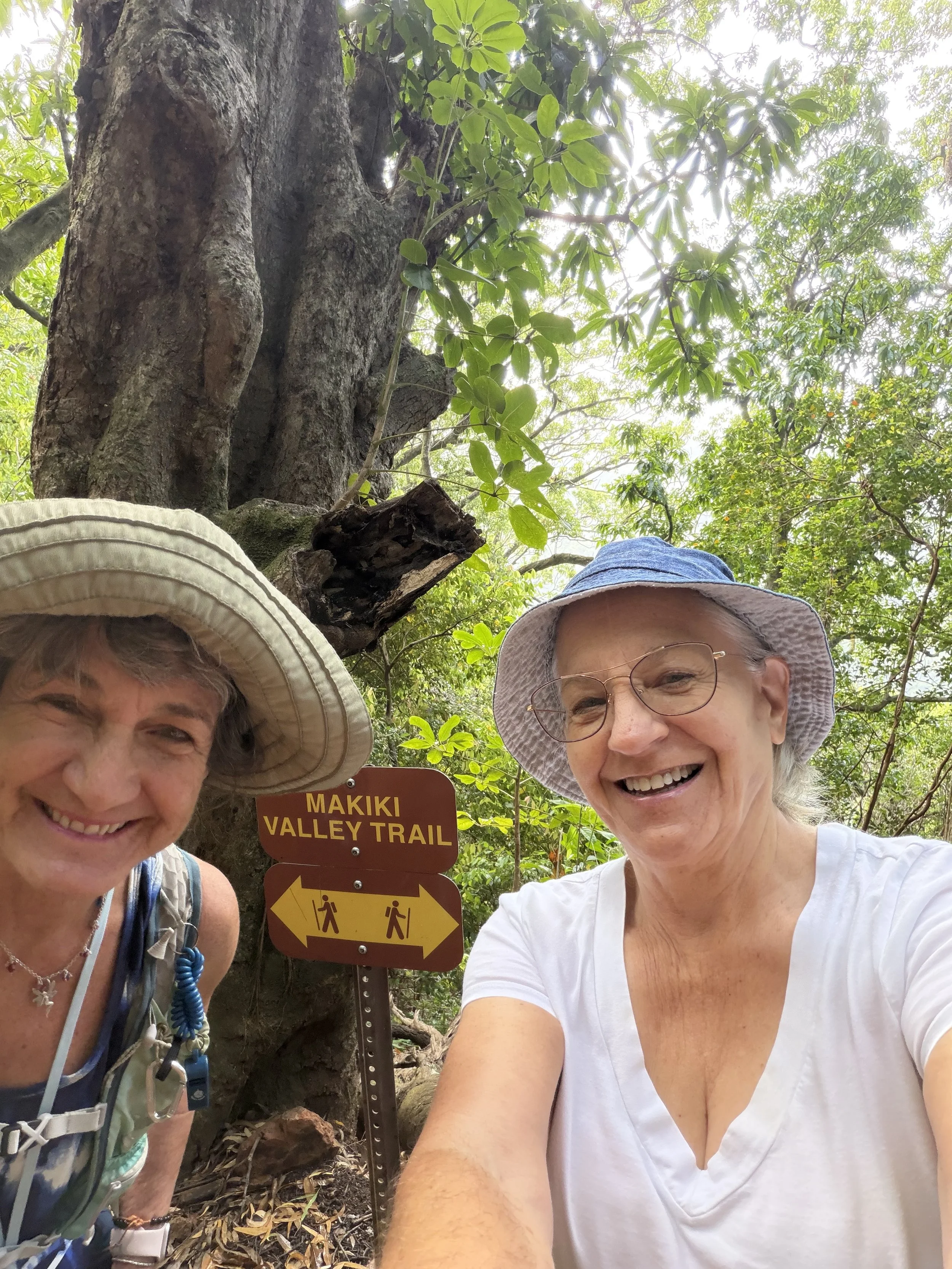 Two women smiling for a selfie in a wooded area at Makiki Valley Trail. One woman is wearing a hat and white shirt, the other is wearing a beige hat and blue scarf. There is a signpost behind them with arrows indicating the trail.