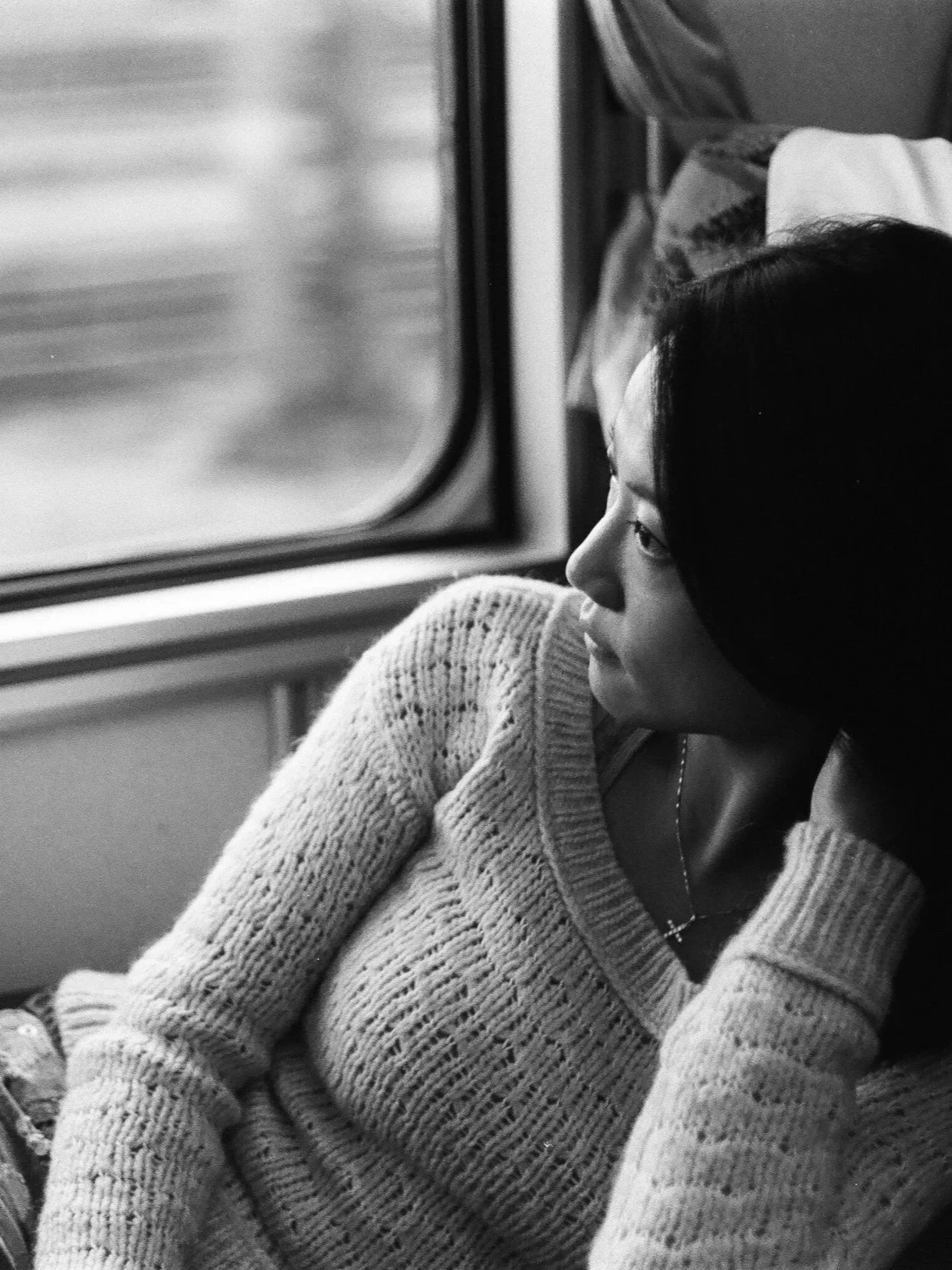 A woman with dark hair wearing a knit sweater and a cross necklace, looking out a train window.