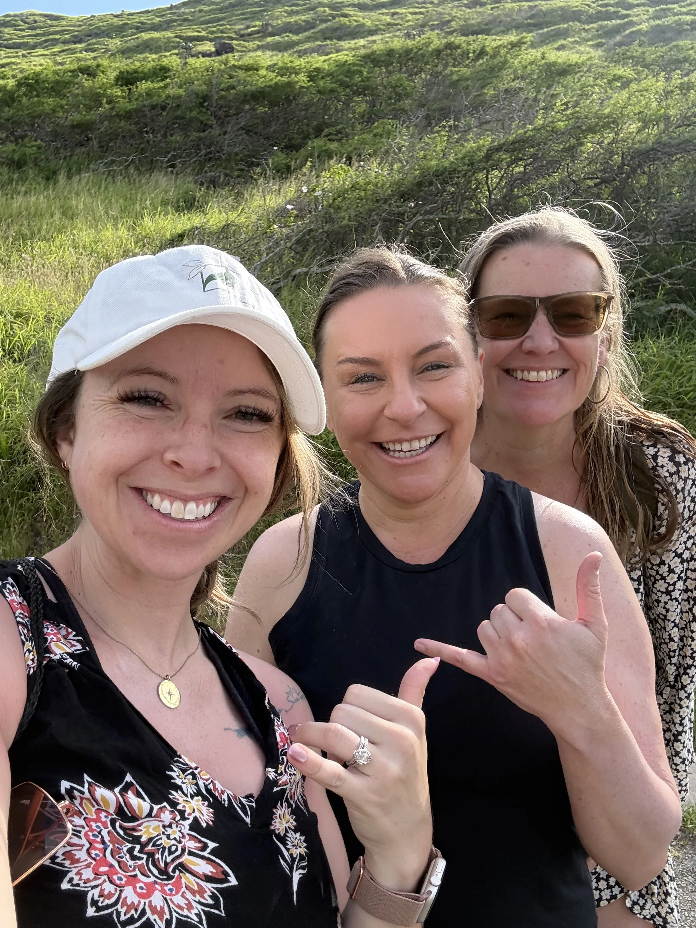 Three women smiling outdoors on a sunny day with greenery and hills in the background, taking a selfie together.