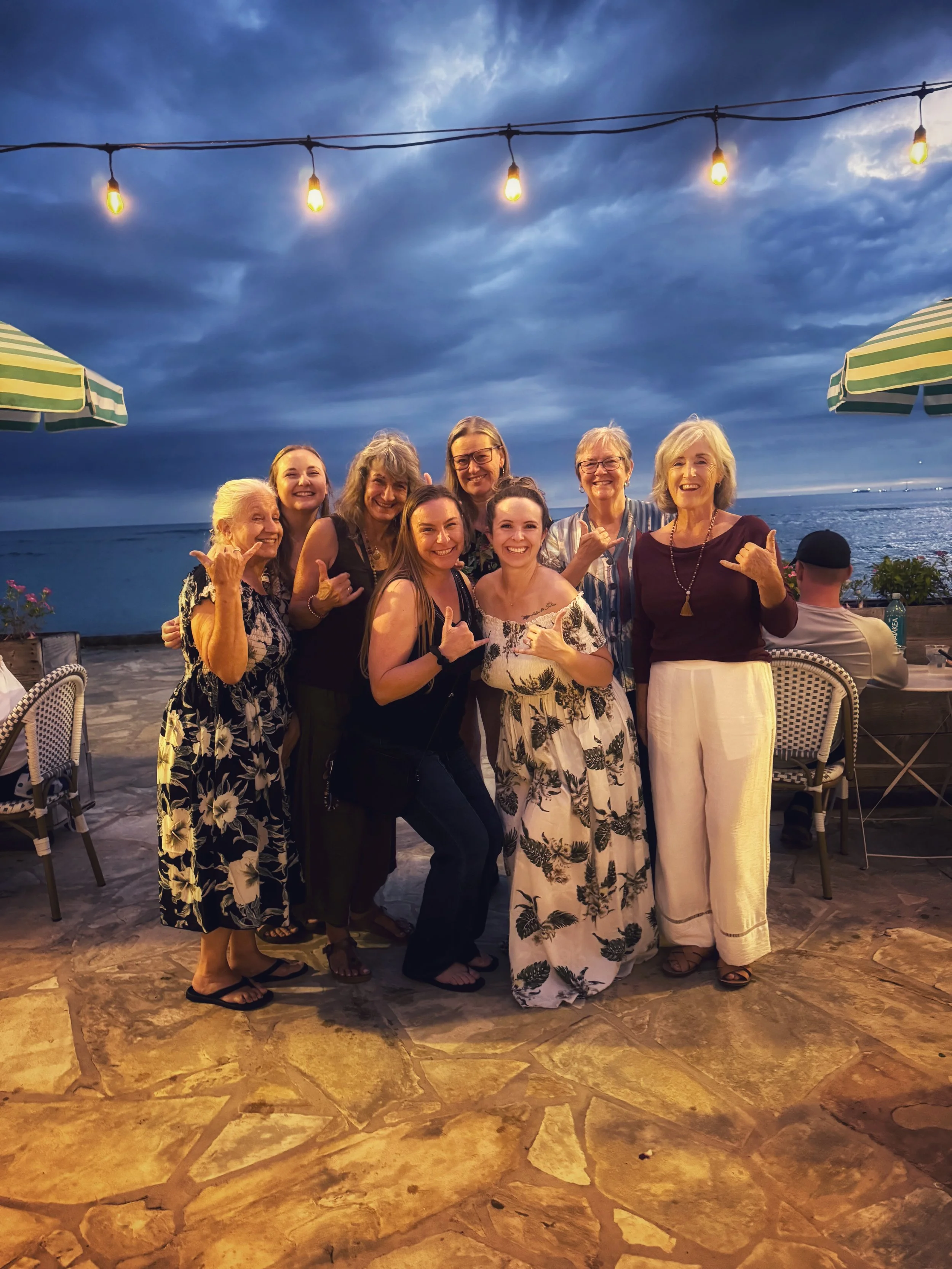 A group of nine women posing together on a patio near the ocean during evening, with a dark cloudy sky overhead and string lights above them, some making hand gestures and smiling.