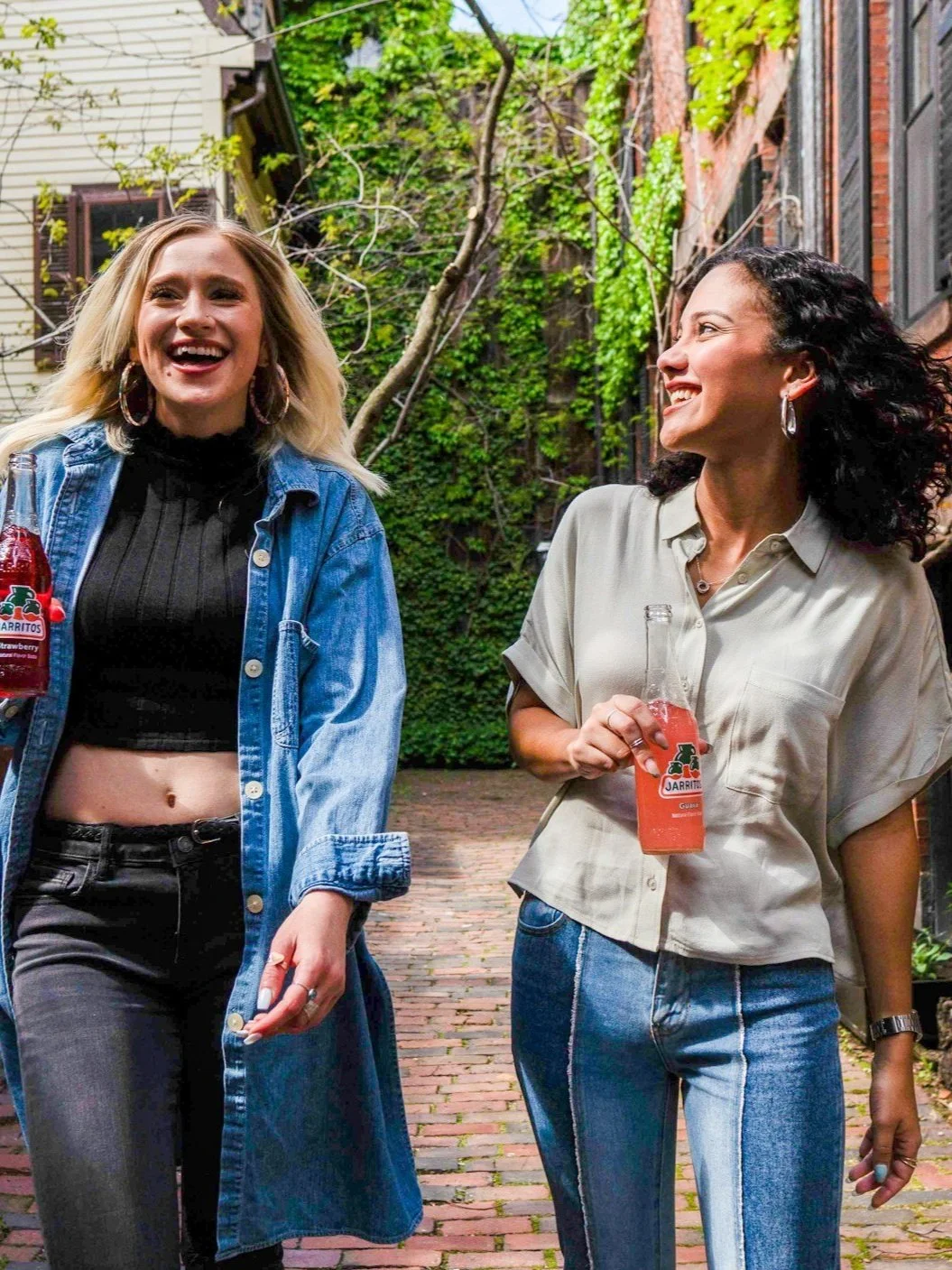 Two women smiling and walking down a brick alleyway, each holding a bottle of Jarritos soda, with green ivy and brick buildings in the background.