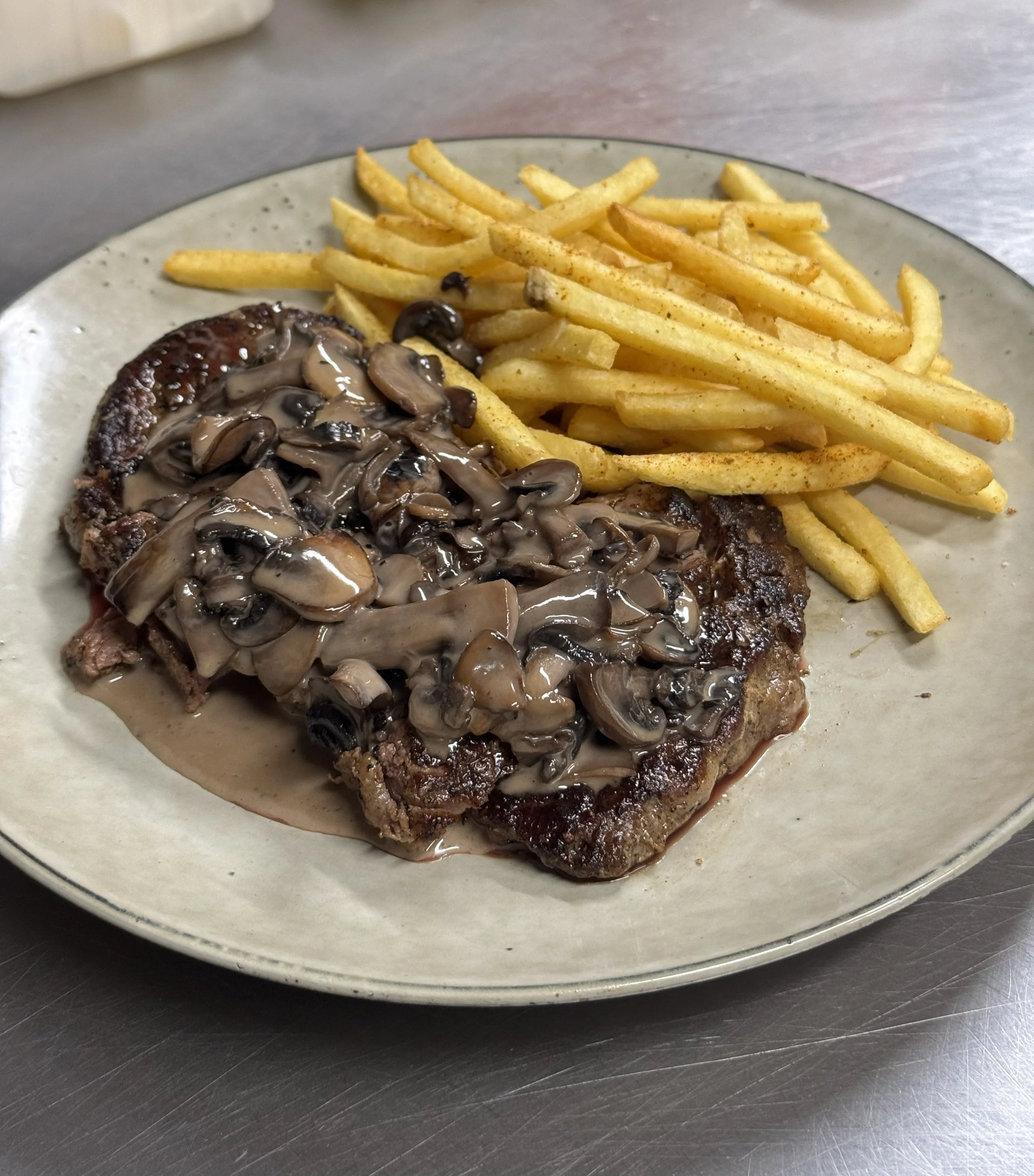 A plate of cooked french fries and a steak topped with sautéed mushrooms and gravy.