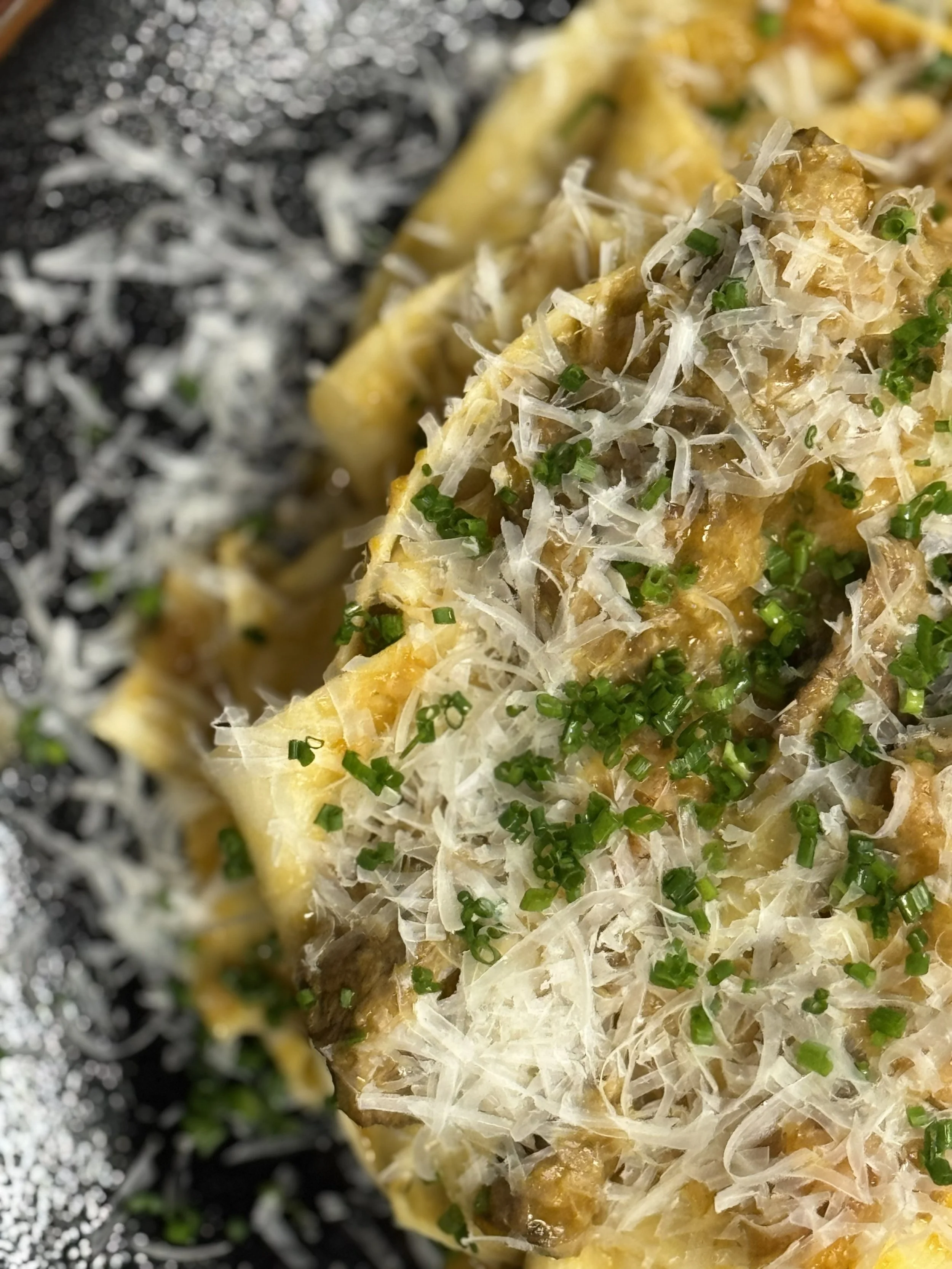 Close-up of a dish with shredded cheese, chopped green herbs, and bite-sized pieces of a creamy, golden-brown food, possibly chicken, on an outdoor grill or griddle.