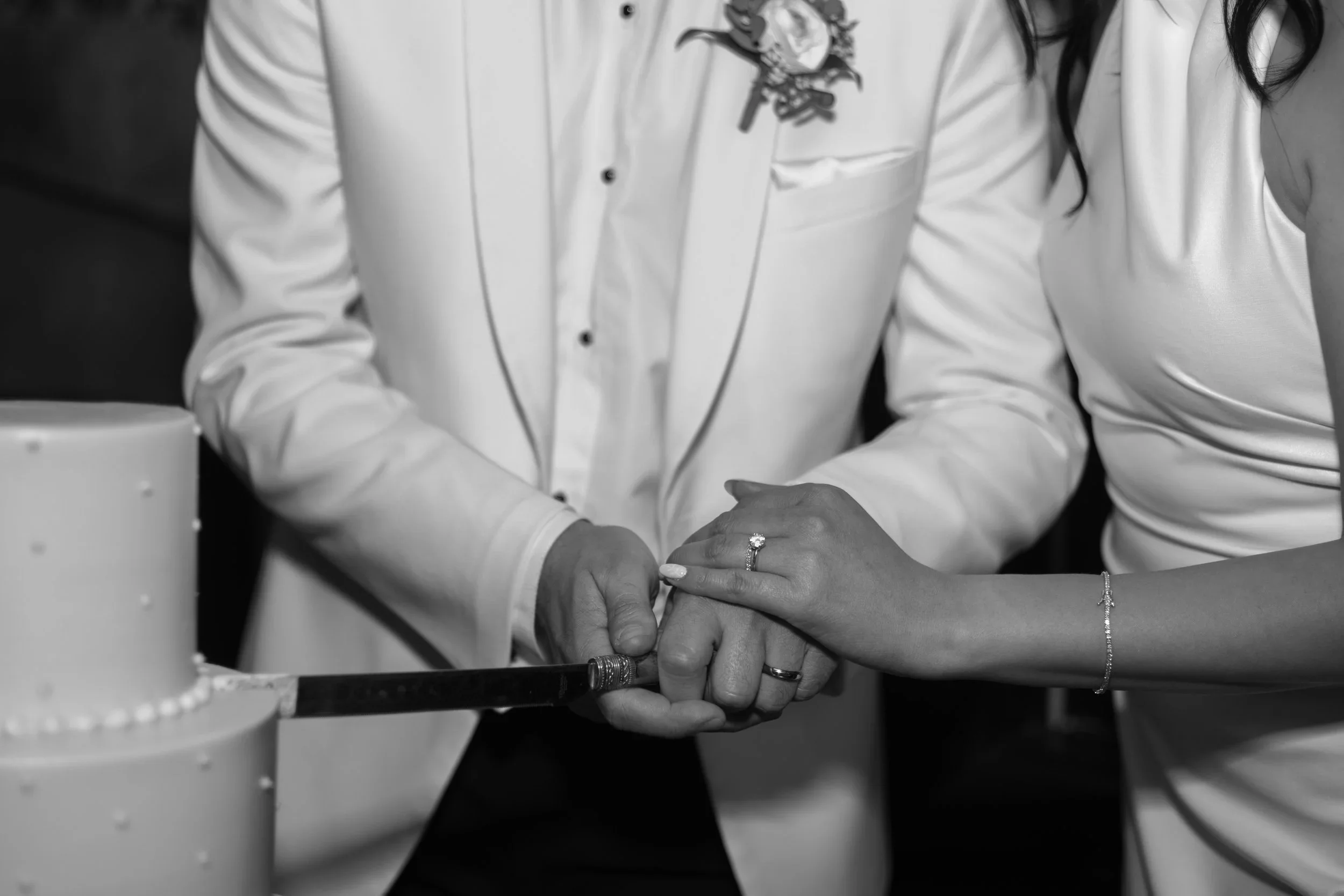Black and white photo of a couple holding hands while cutting a wedding cake. The woman wears an engagement ring and a bracelet, and the man wears a ring and a white shirt with embroidery.