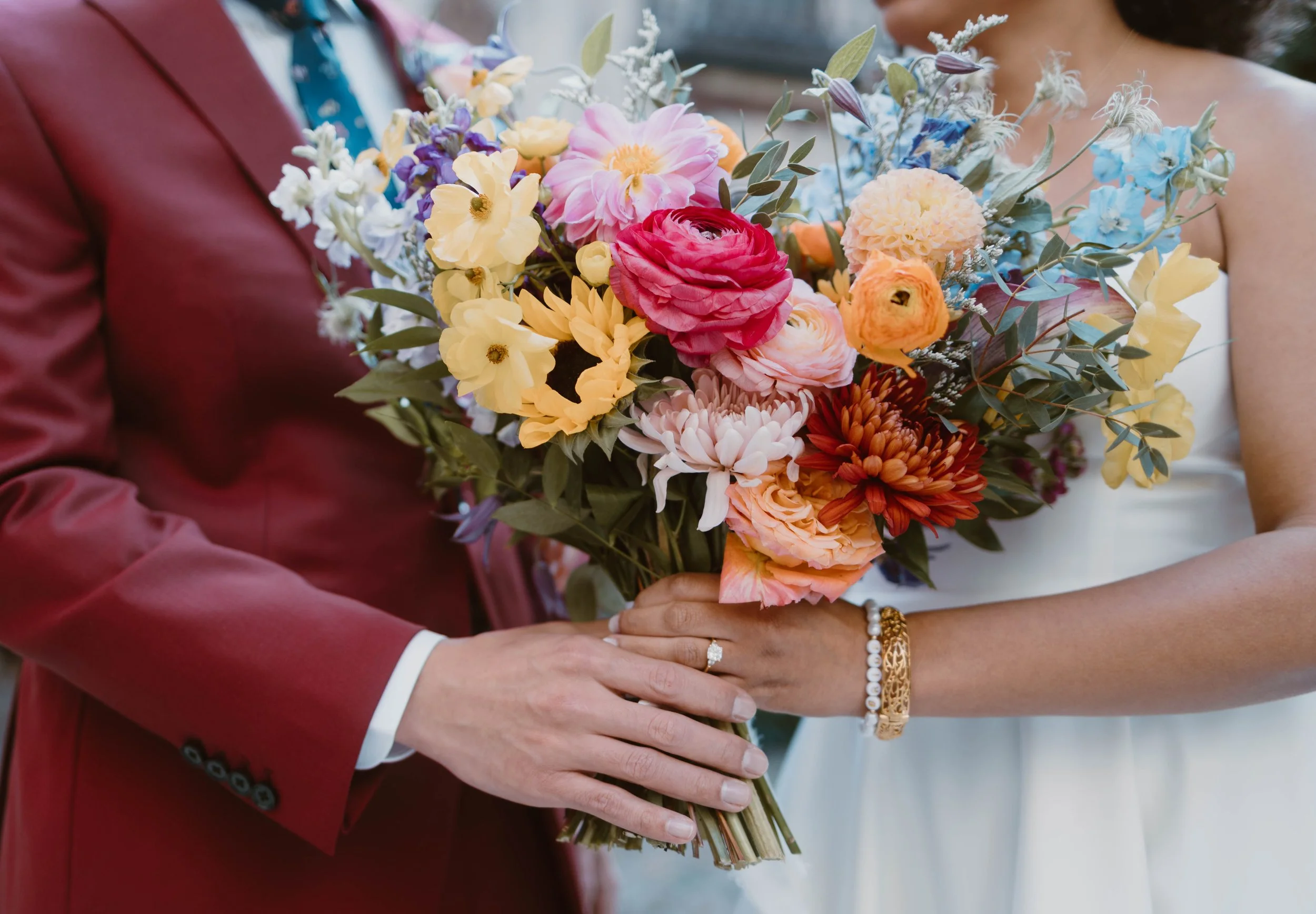 Close-up of a bride and groom holding a vibrant bouquet of flowers during their wedding. The bride is wearing a white dress and jewelry, and the groom is in a red suit with a tie.