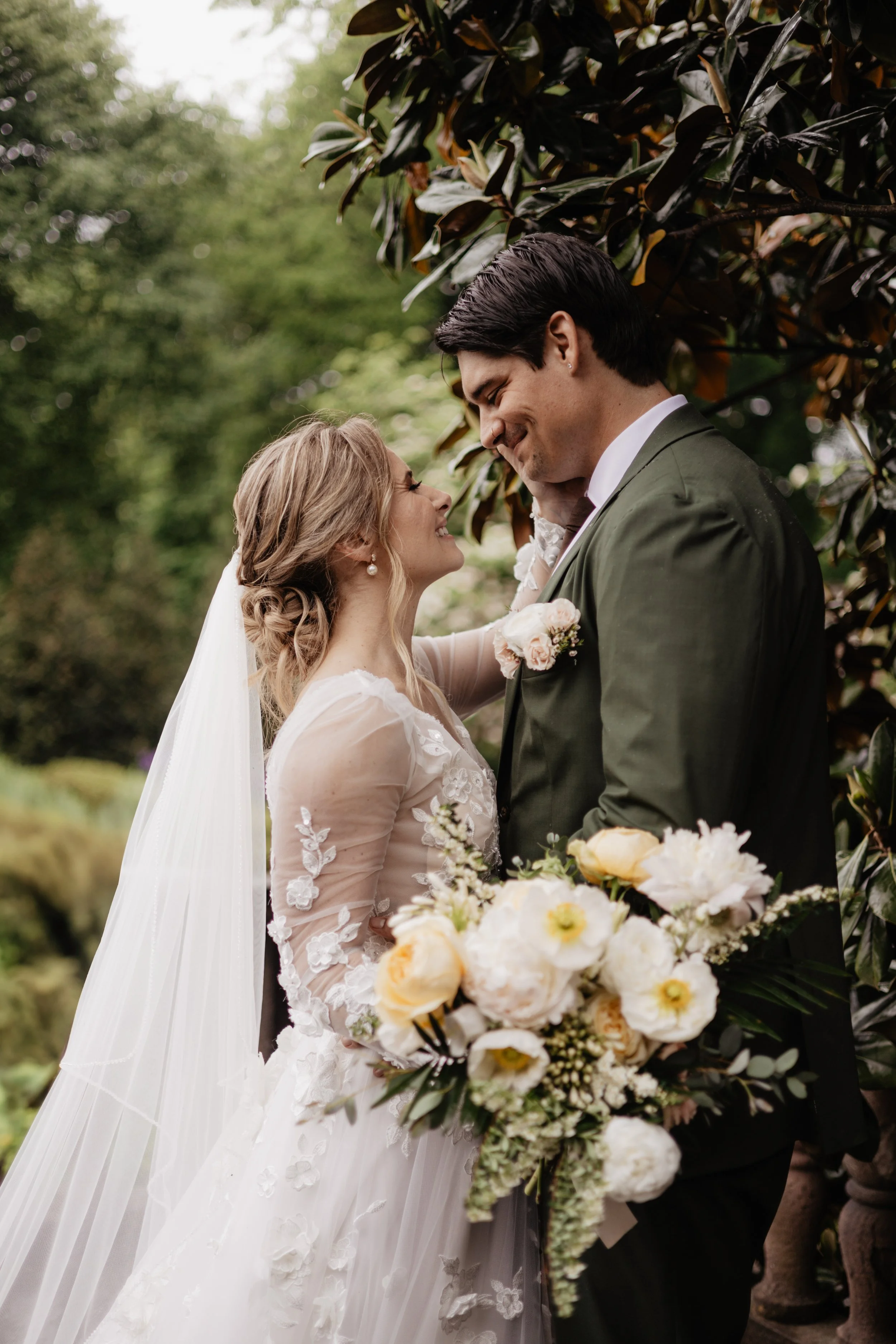 Bride and groom sharing a tender moment outdoors, with the bride holding a bouquet of white and yellow flowers, both smiling and leaning close to each other.