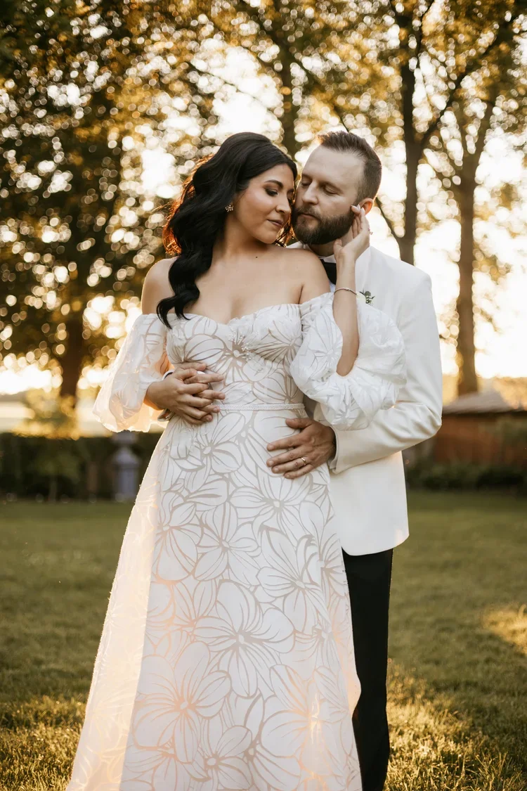 A couple on their wedding day, standing outdoors during sunset, embracing lovingly with trees and soft sunlight in the background.