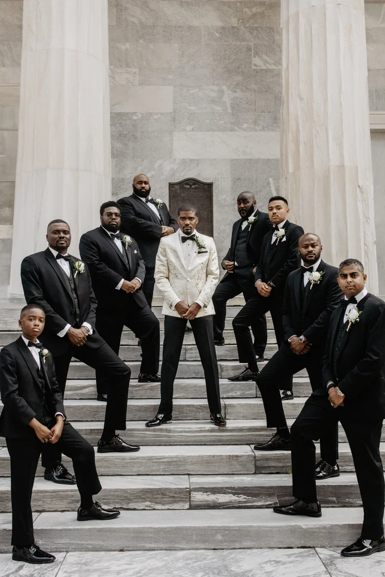 A groom in a white tuxedo jacket and bow tie standing with his groomsmen in black suits on marble stairs.