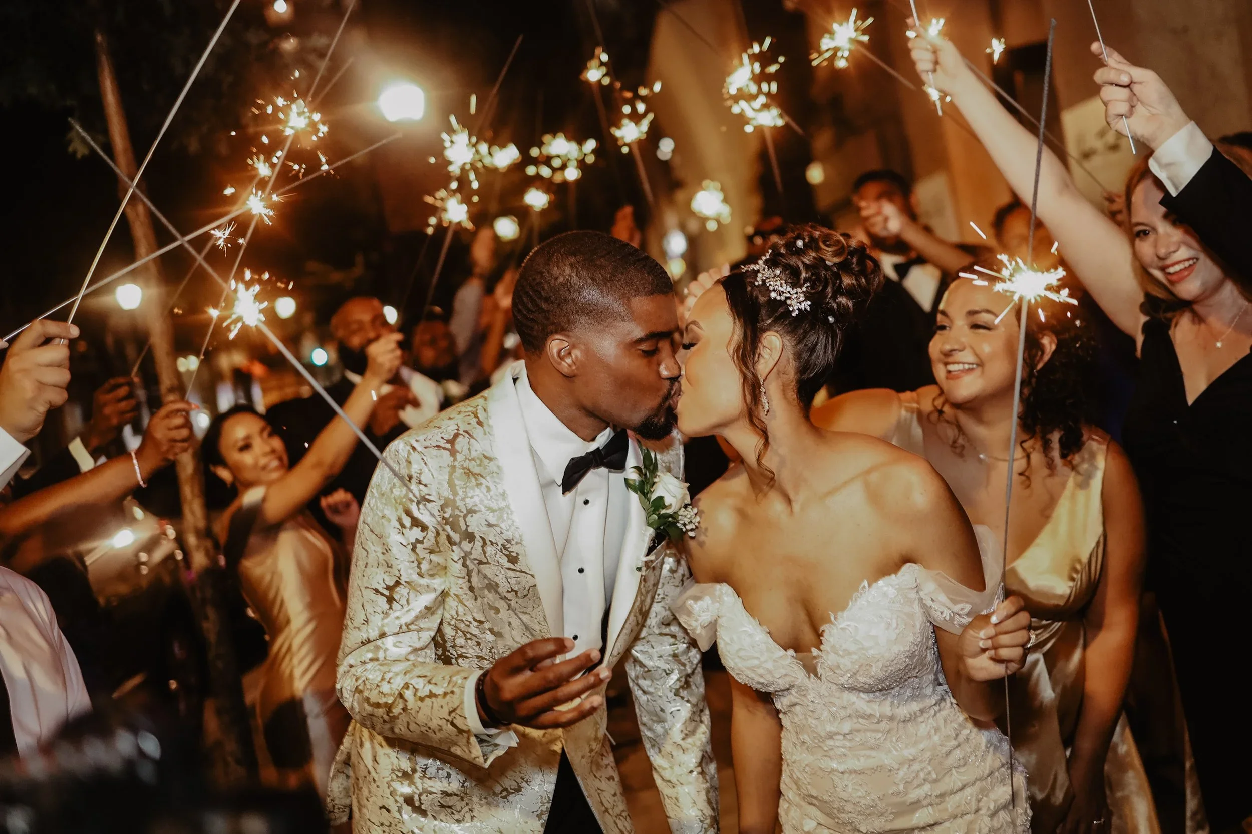 A newlywed couple shares a kiss at their wedding celebration surrounded by friends and family holding sparklers at night.