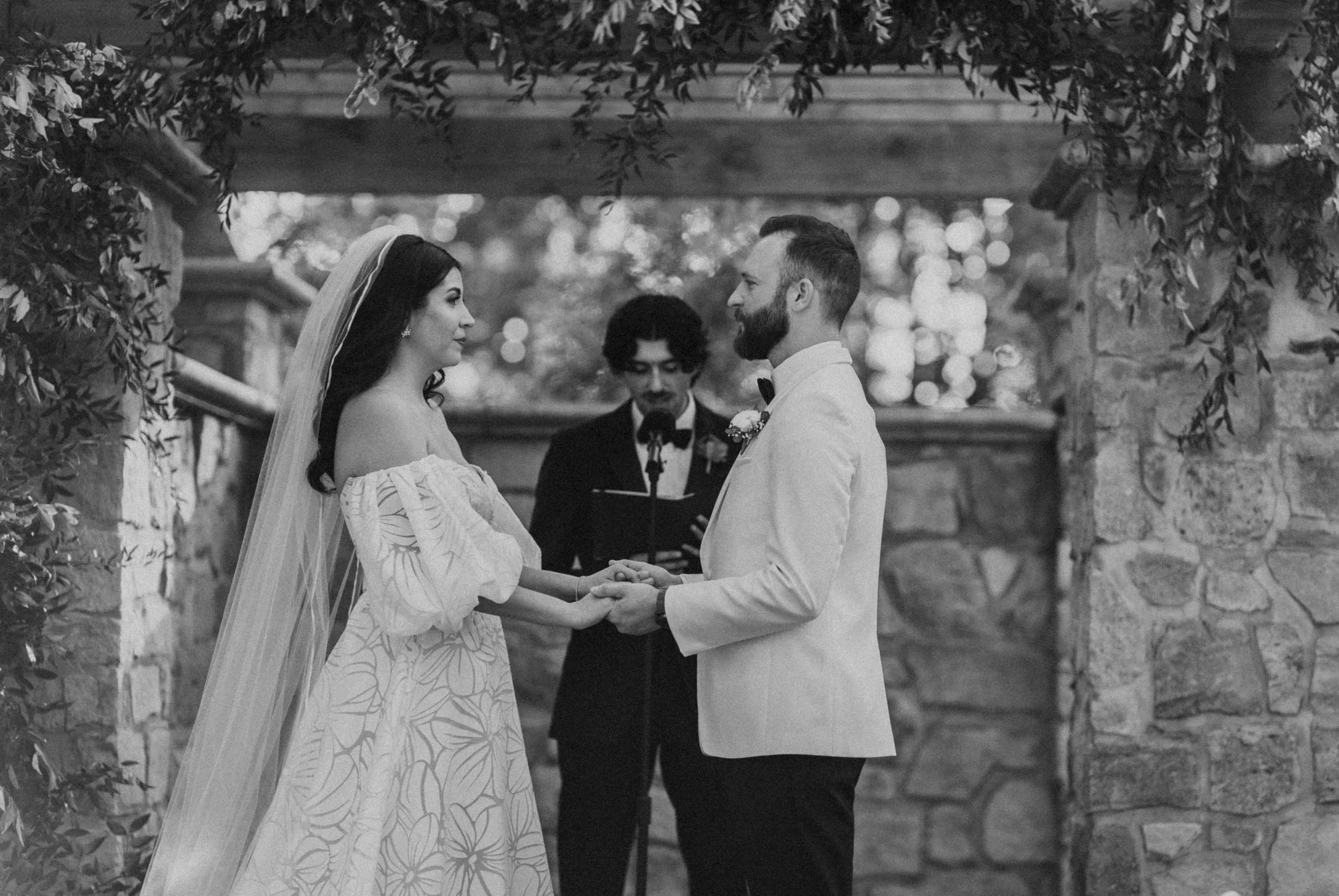 Black and white photo of a bride and groom holding hands during their wedding ceremony outside, with a person officiating in the background holding a book.