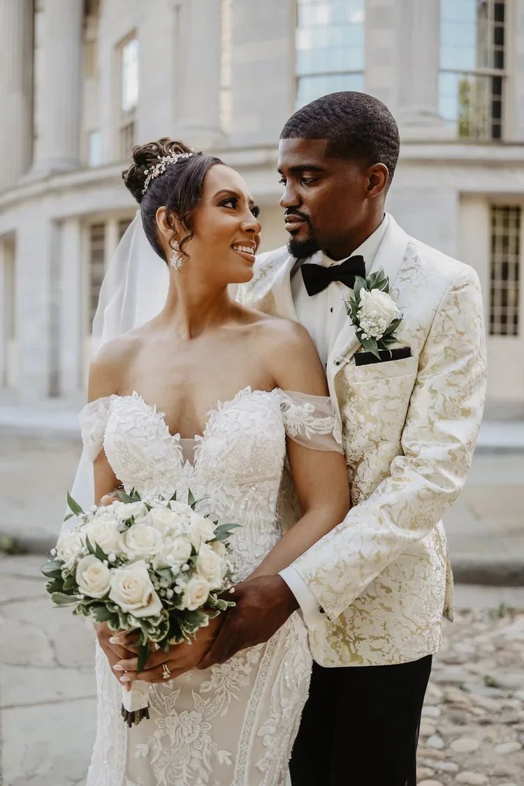 A bride and groom in wedding attire standing outdoors in front of a historic building, holding a bouquet of white roses, looking at each other lovingly.