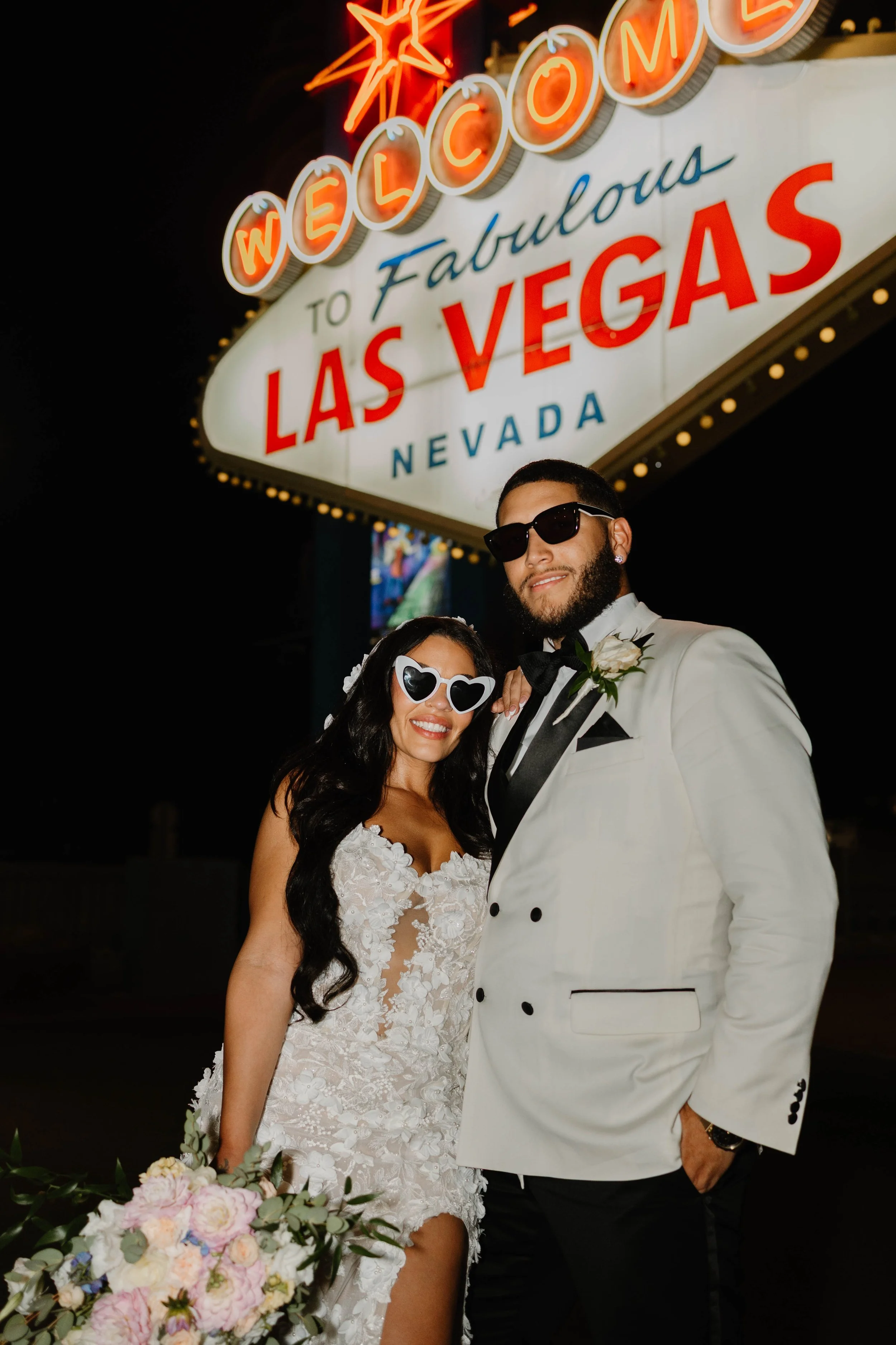 A newlywed couple dressed in wedding attire stands in front of the illuminated 'Welcome to Fabulous Las Vegas Nevada' sign at night. The bride wears a white lace dress and sunglasses, holding a bouquet of flowers, while the groom wears a white tuxedo