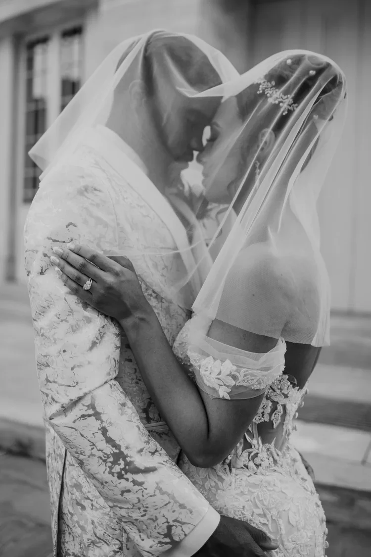 A black-and-white photo of a bride and groom with their foreheads touching, both wearing wedding attire and veils, embracing each other closely.