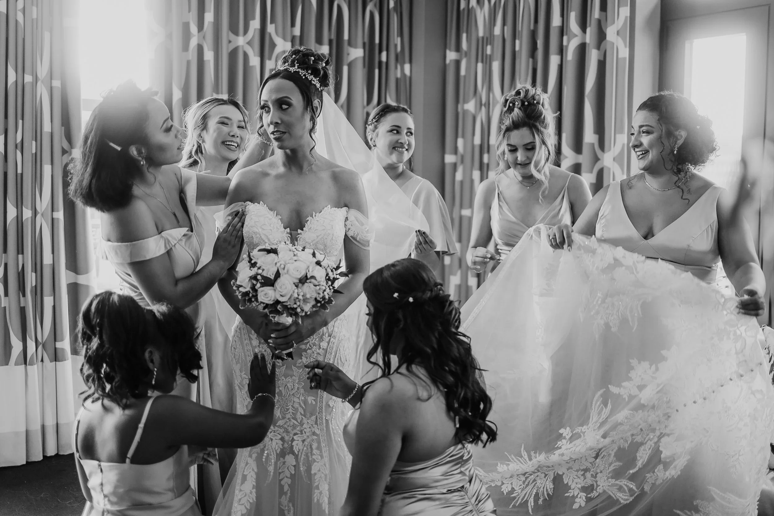 Black and white photo of a bride with her bridesmaids and young flower girls, preparing for the wedding, with the bride holding a bouquet of flowers.