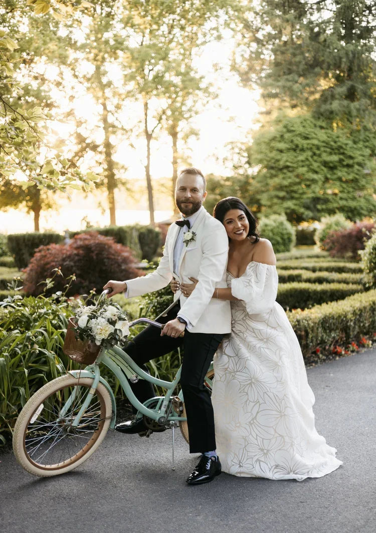 A newlywed couple, dressed in wedding attire, posing outdoors at sunset. The groom is sitting on a vintage bicycle decorated with flowers, and the bride is standing beside him, smiling and hugging him. The setting features lush greenery and a peacefu