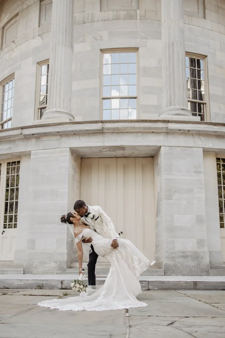 Couple in wedding attire sharing a kiss outside a large, classical-style building with columns and tall windows.