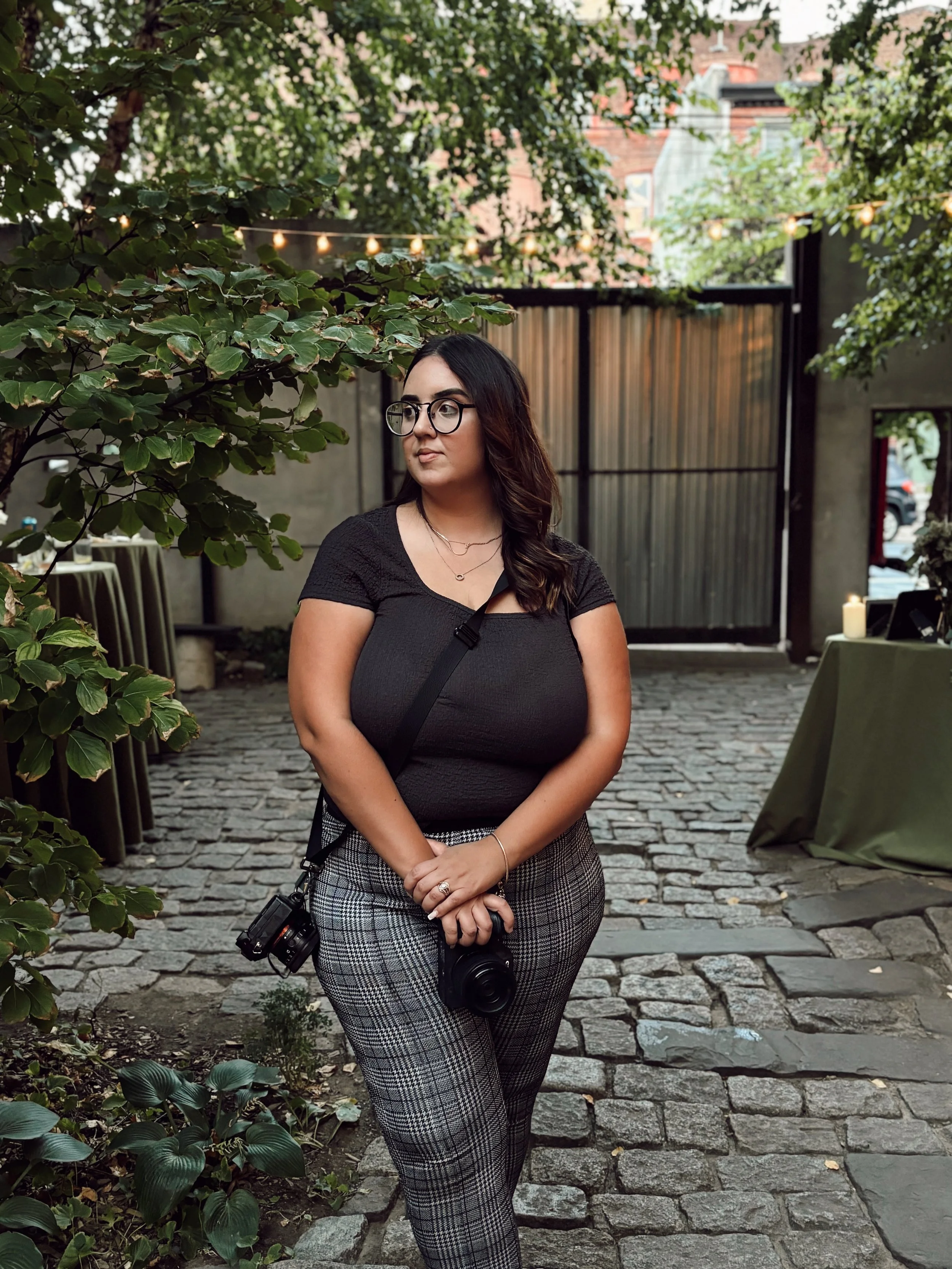 A woman with glasses and a camera stands outdoors on a cobblestone path, surrounded by green trees and string lights.