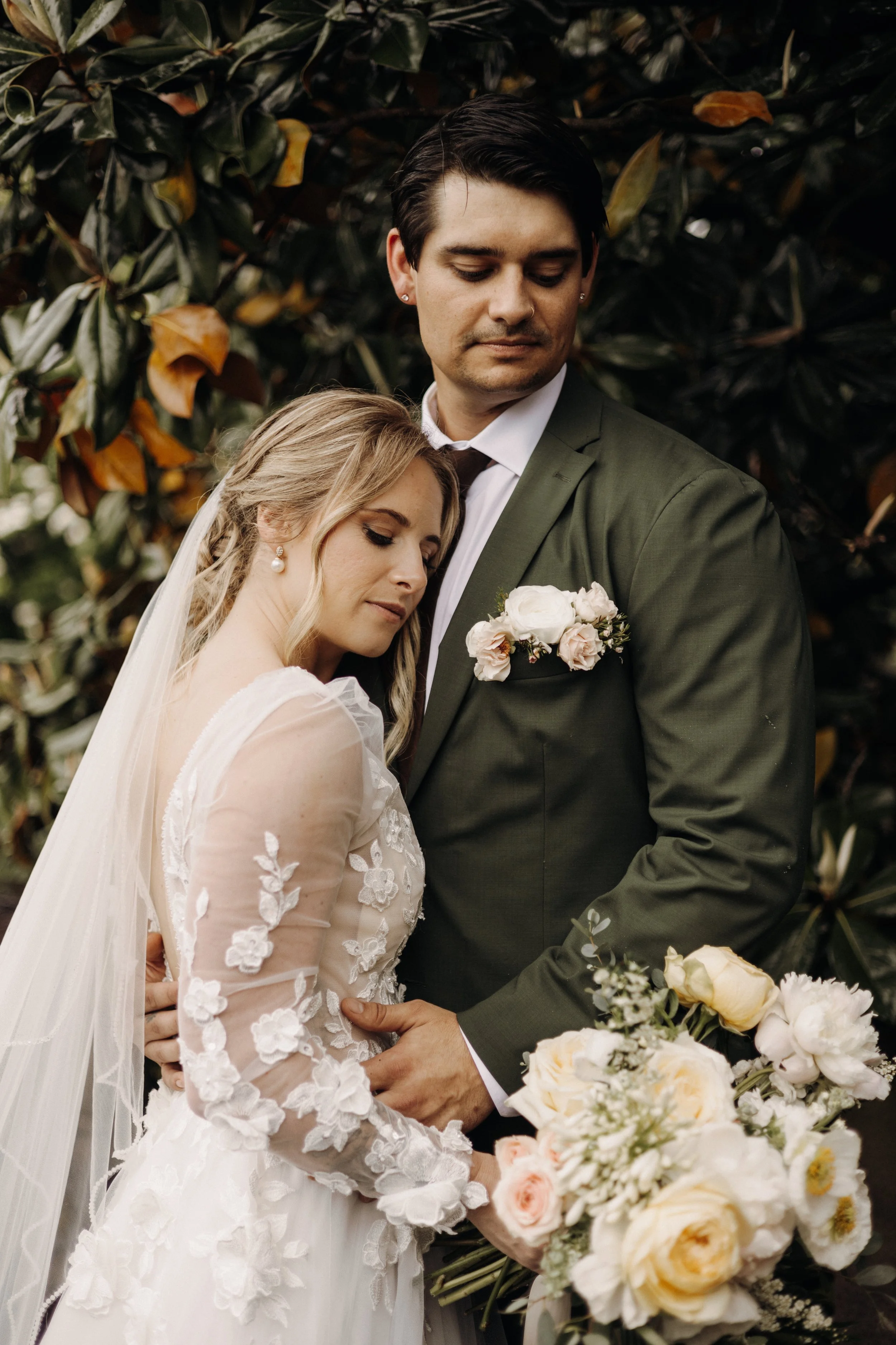 A bride and groom standing close together outdoors, the bride resting her head on the groom's chest. The bride is in a white wedding dress with floral embroidery and a veil, and she holds a bouquet of white and pale pink flowers. The groom wears an o