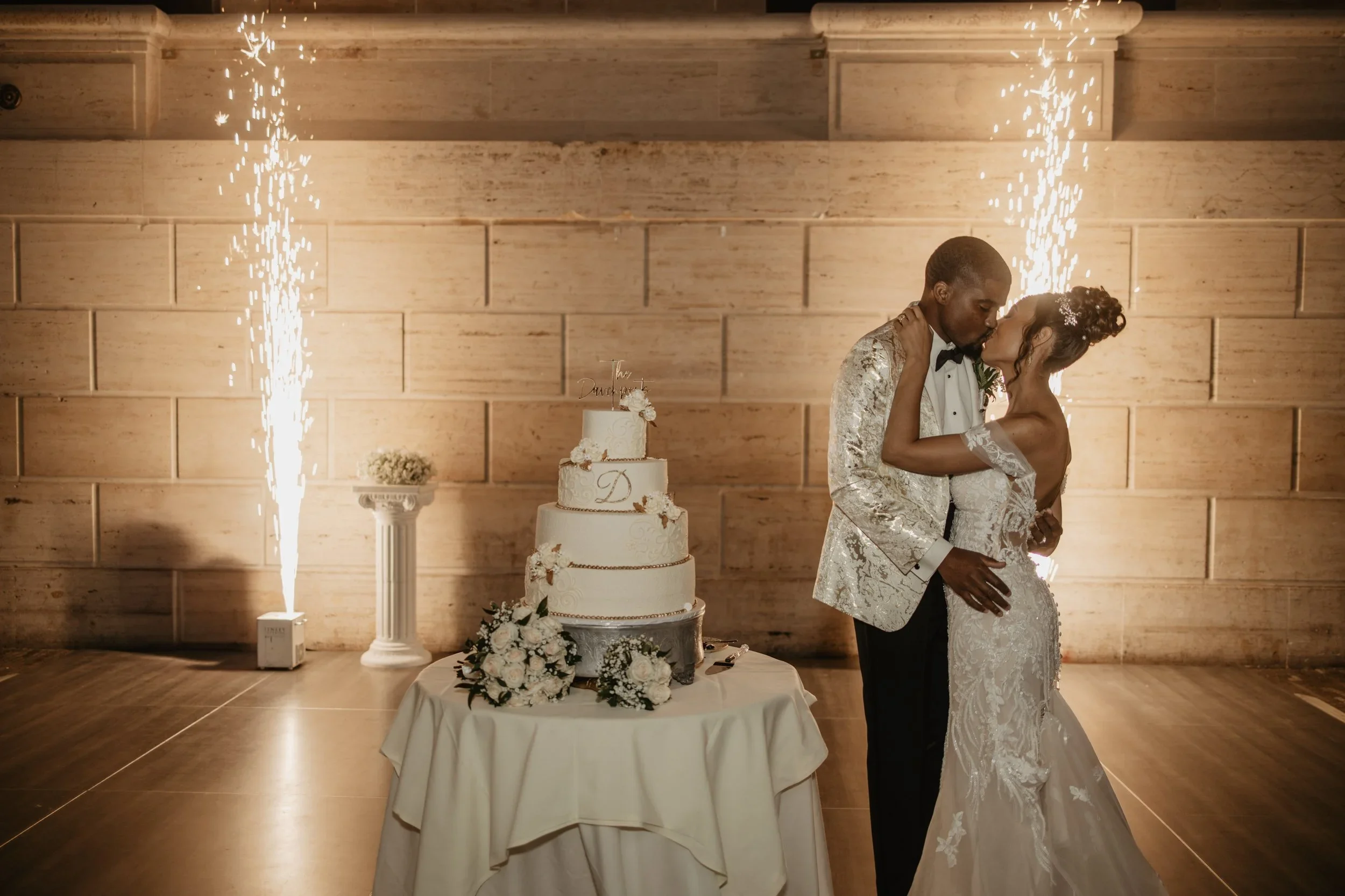 A newlywed couple sharing a kiss at their wedding reception, with sparklers on either side, a wedding cake on a table, and a brick wall background.